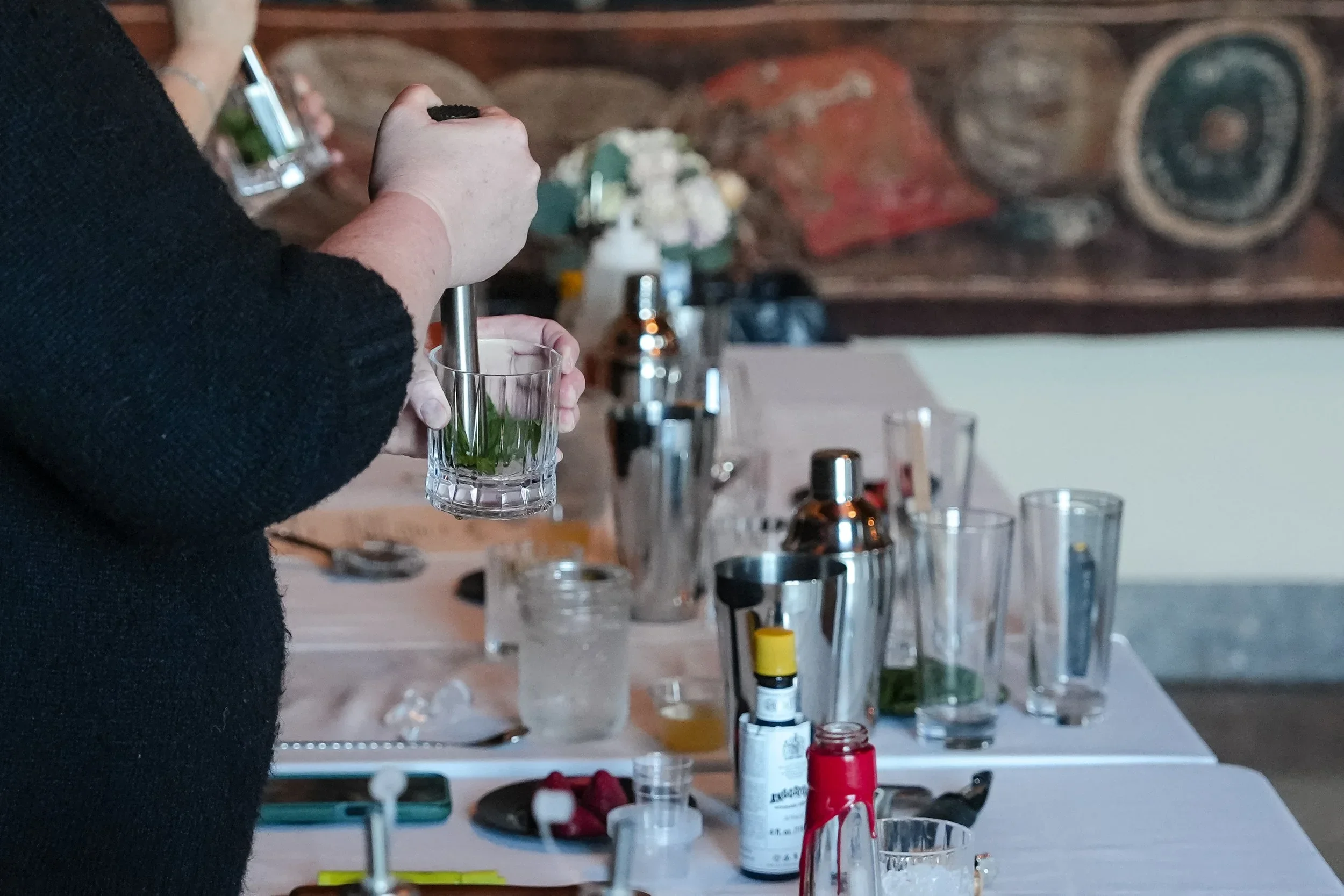 Person wearing black shirt and gloves using a pestle to crush green herbs inside a glass. The background shows a tabletop with various bottles and containers, and a rustic wooden wall with a round decorative item.
