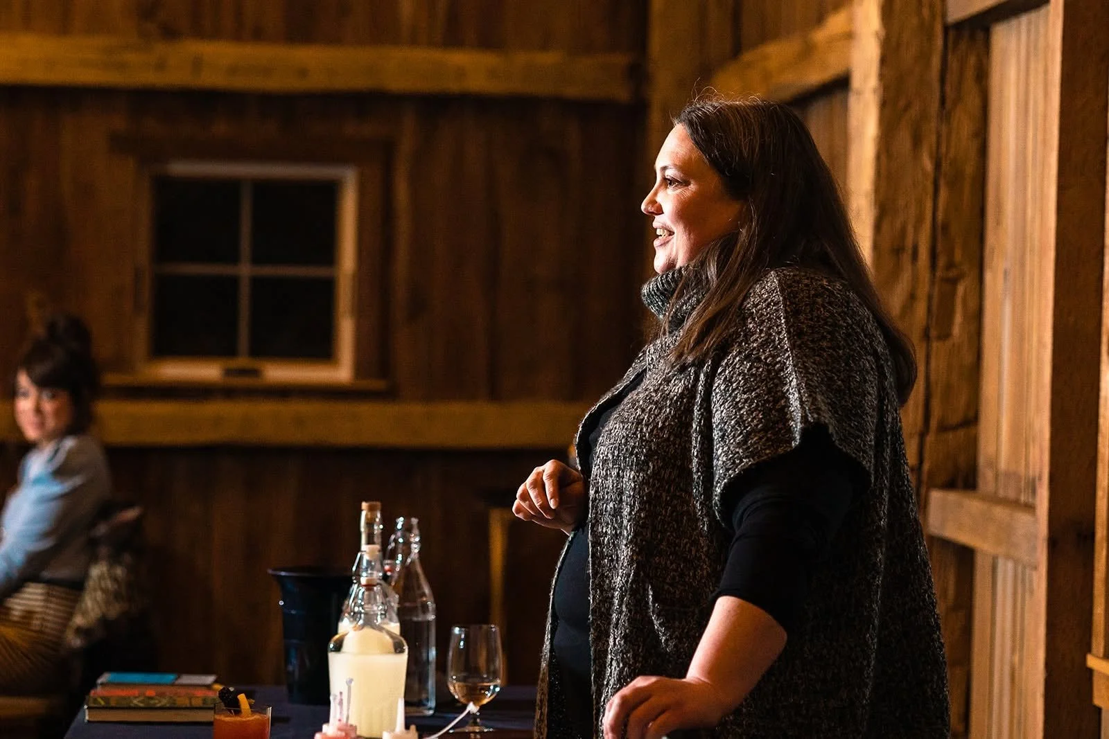 A woman with long dark hair smiling, standing in a rustic wooden room, with a second woman sitting in the background. There are bottles, a book, and candles on a table in front of her.