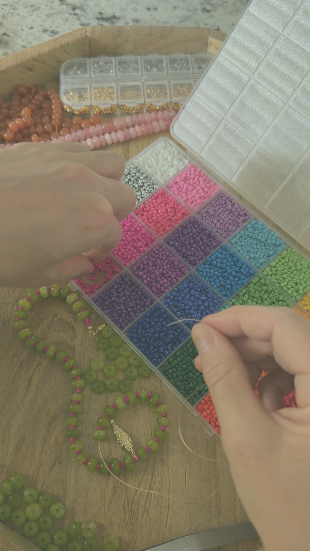 Hands working with small colorful beads, organizing them in a plastic compartment box for jewelry making or crafting, on a wooden surface.