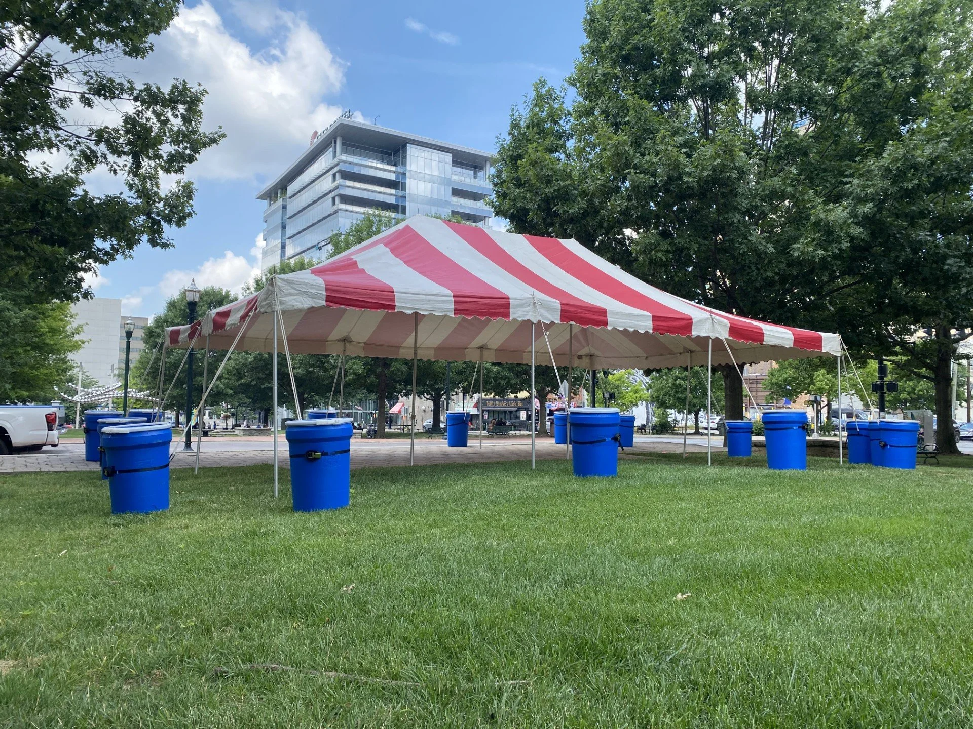 A park with green grass, large trees, and a red and white striped canopy tent. Several large blue buckets are arranged around the tent. In the background, there is a multi-story glass building and some street lamps.