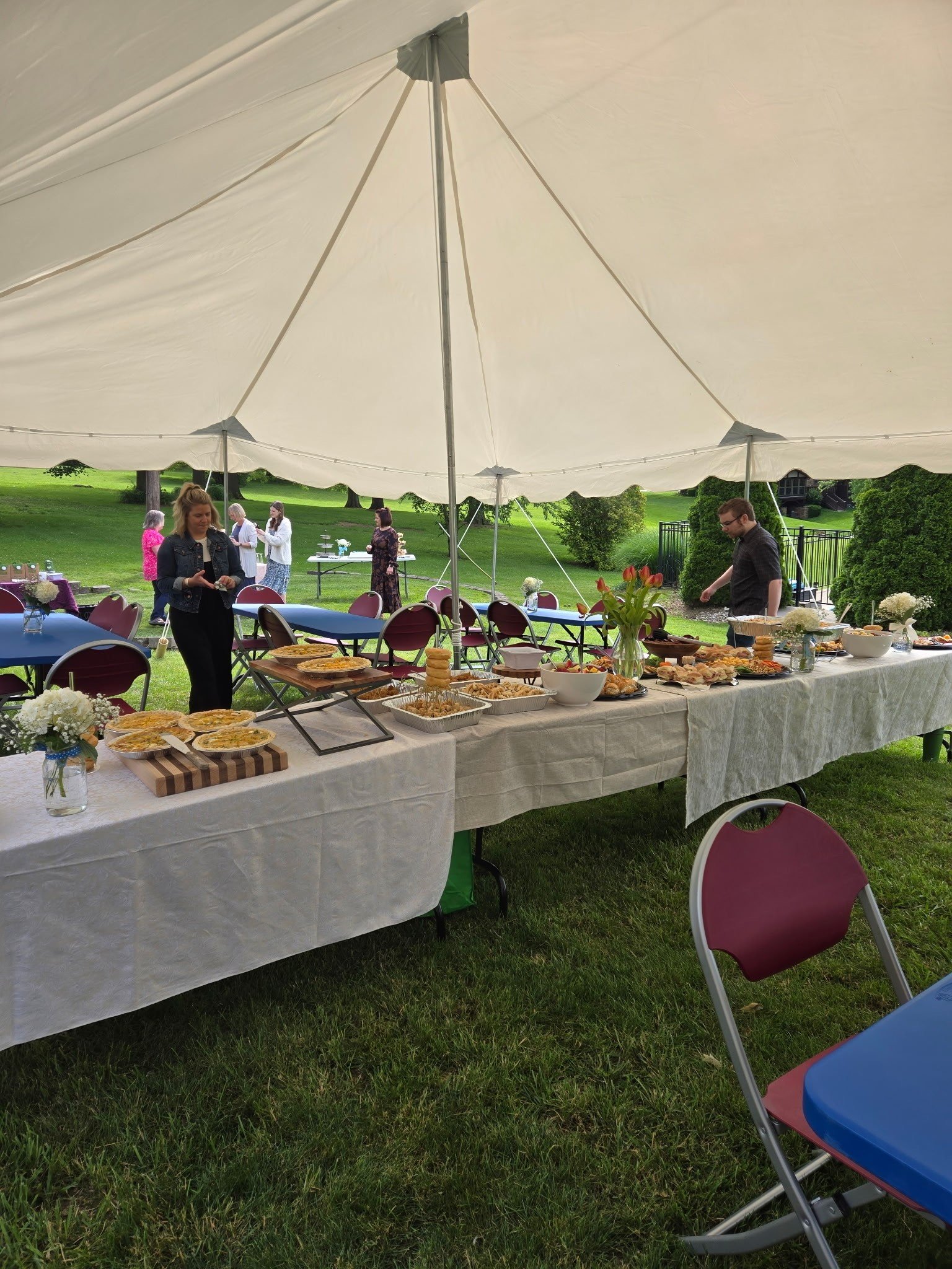 Outdoor gathering under a large white 20x30ft tent with a buffet table filled with various foods, surrounded by red and blue chairs, on a grassy lawn with trees in the background in Frankfort, Kentucky
