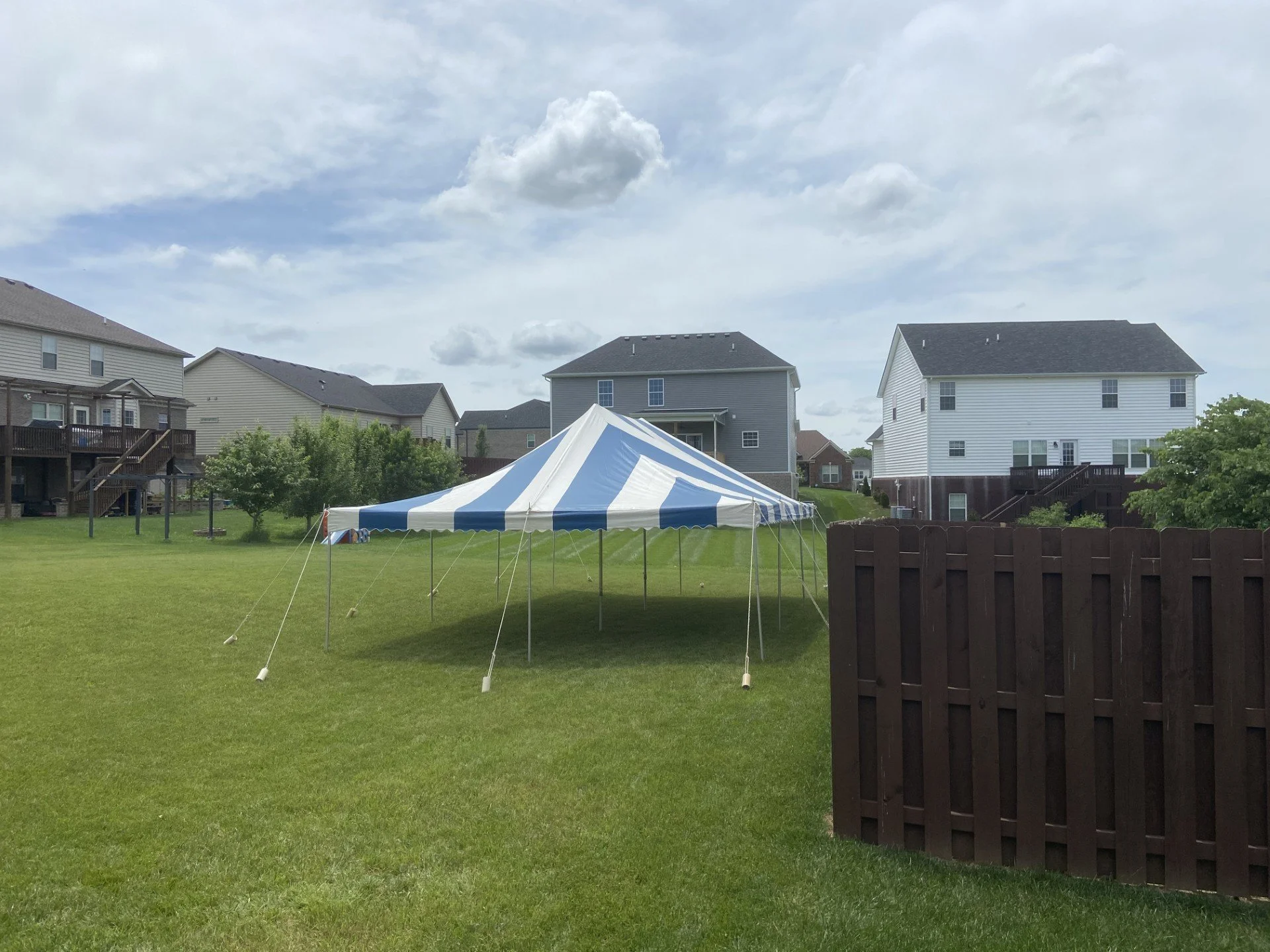 A backyard with green grass, set up with a white and blue striped party tent, surrounded by houses and trees under a partly cloudy sky.