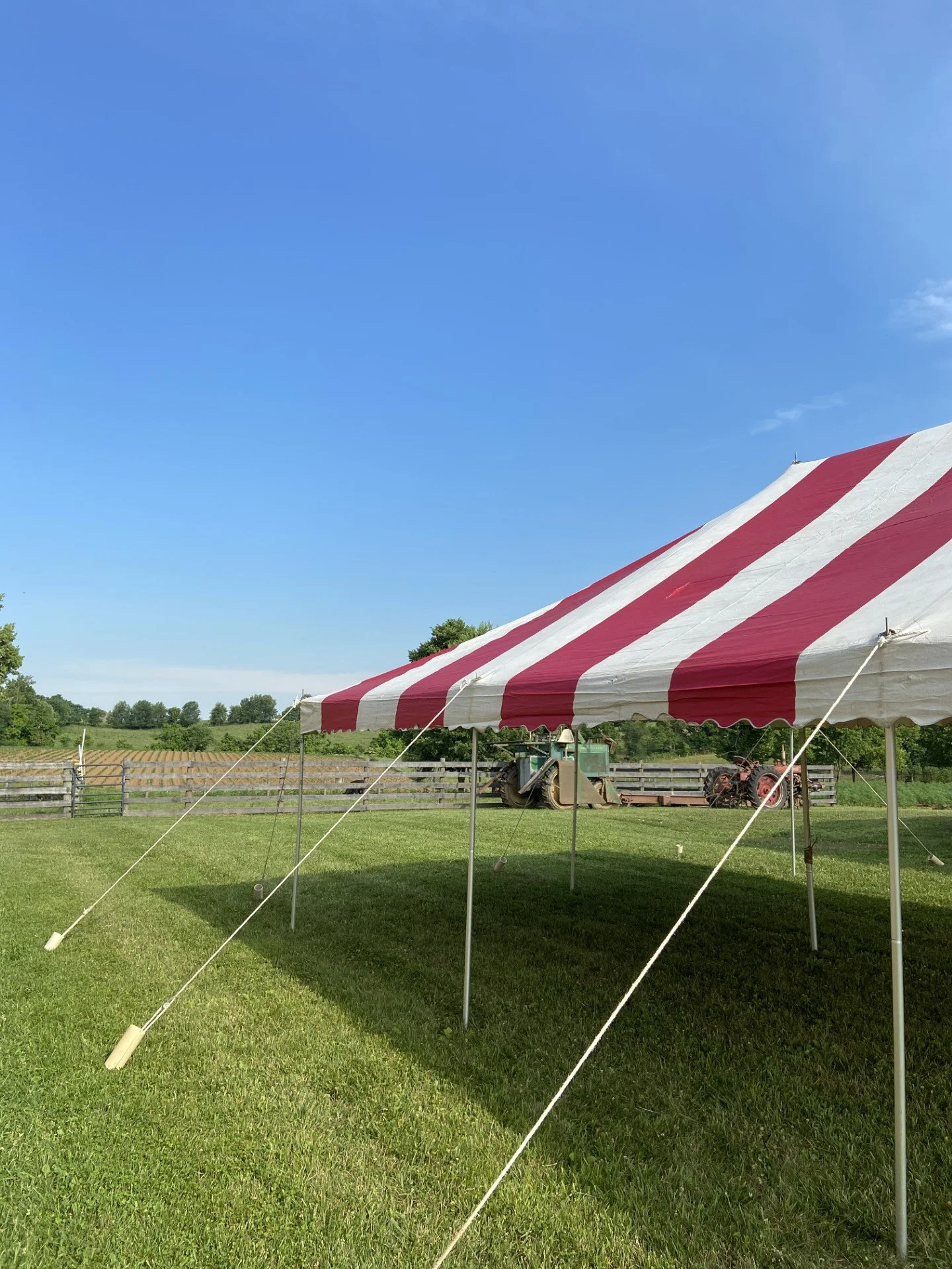 A red and white striped canopy tent set up on a grassy field with a clear blue sky overhead. In the background, there are farm equipment and a wooden fence with a scenic rural landscape of trees and fields.
