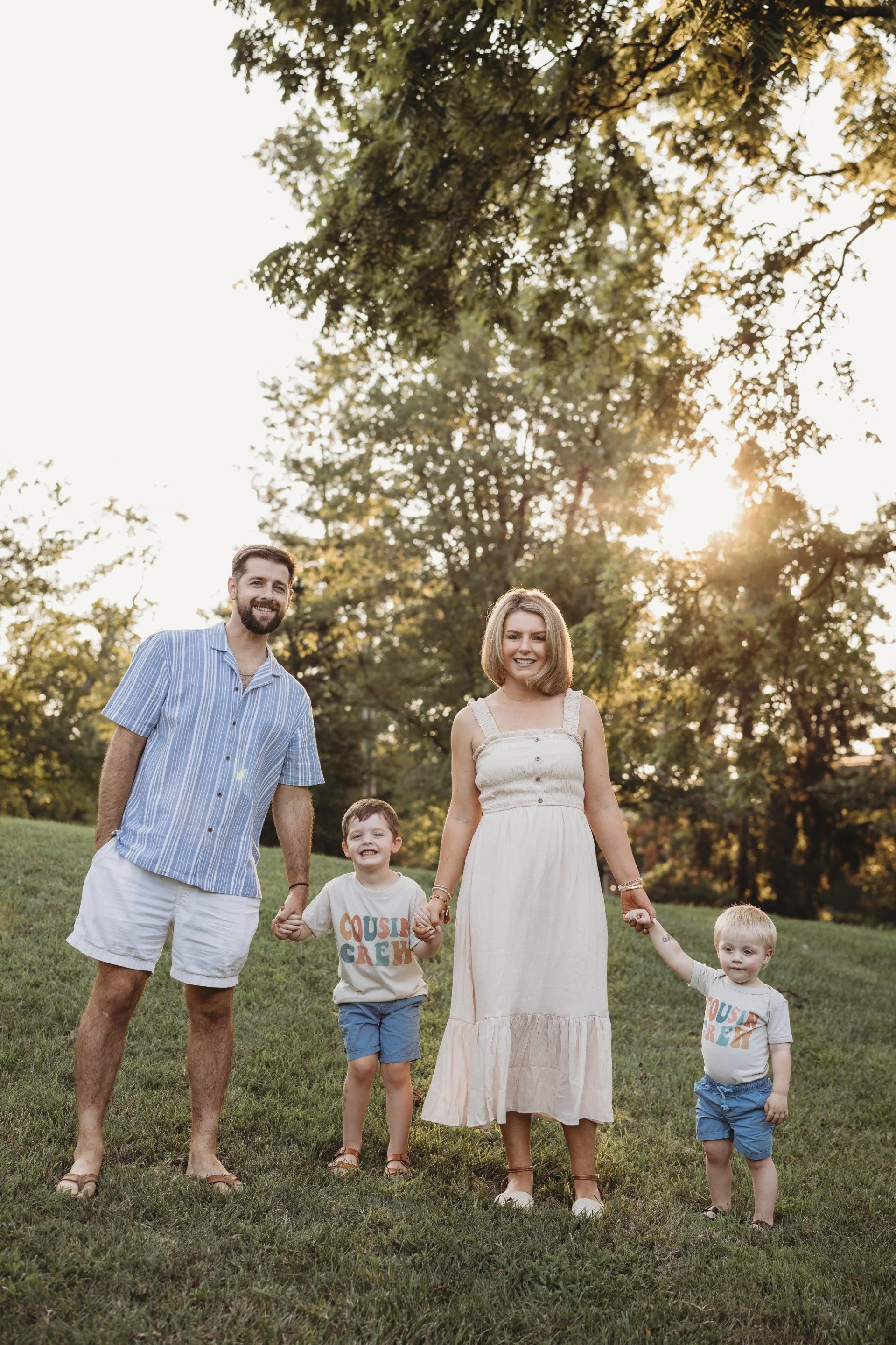 A family of five, including two adults and three children, holding hands and walking outdoors in a grassy park during sunset with trees in the background.