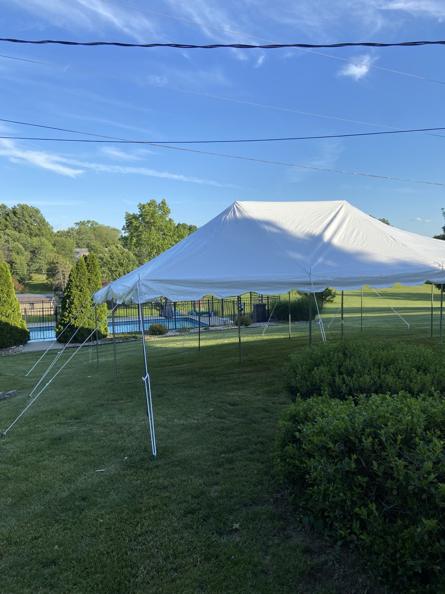 Large white event tent set up on a grassy backyard with trees and a swimming pool in the background, under a blue sky with some clouds.