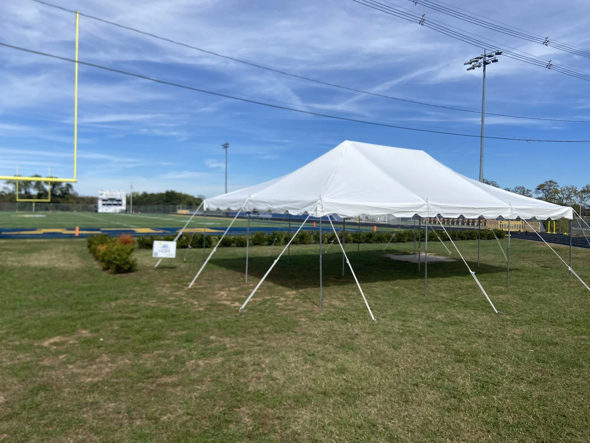 A sports field with a large white event tent set up on the grass near a running track and goalposts, with utility poles and wires overhead under a partly cloudy sky.