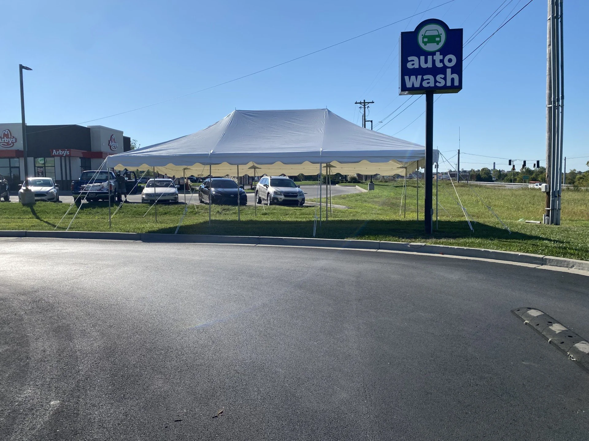 Parking lot with a white tent, several parked cars, a fast food restaurant Arby's in the background, and a blue sign for an auto wash.