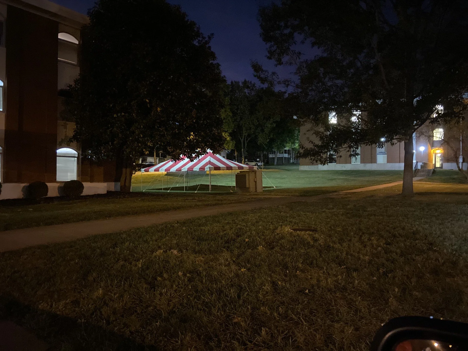 Nighttime scene of a grassy area with large trees, a red and white striped tent, and apartment buildings in the background. Lights are seen in some windows, and the sky is dark.