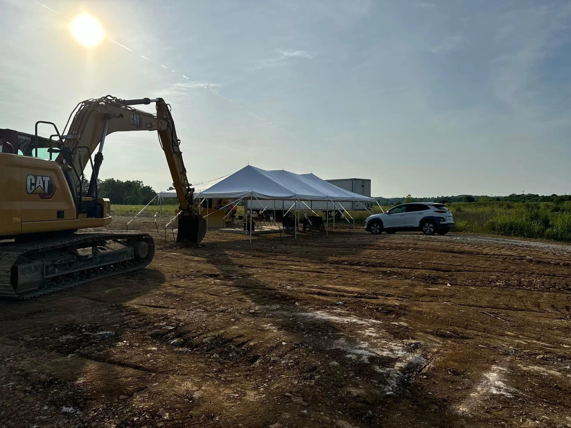 Construction site with a yellow CAT excavator, a white tent, and a white car on dirt ground under a bright sun in a rural area.