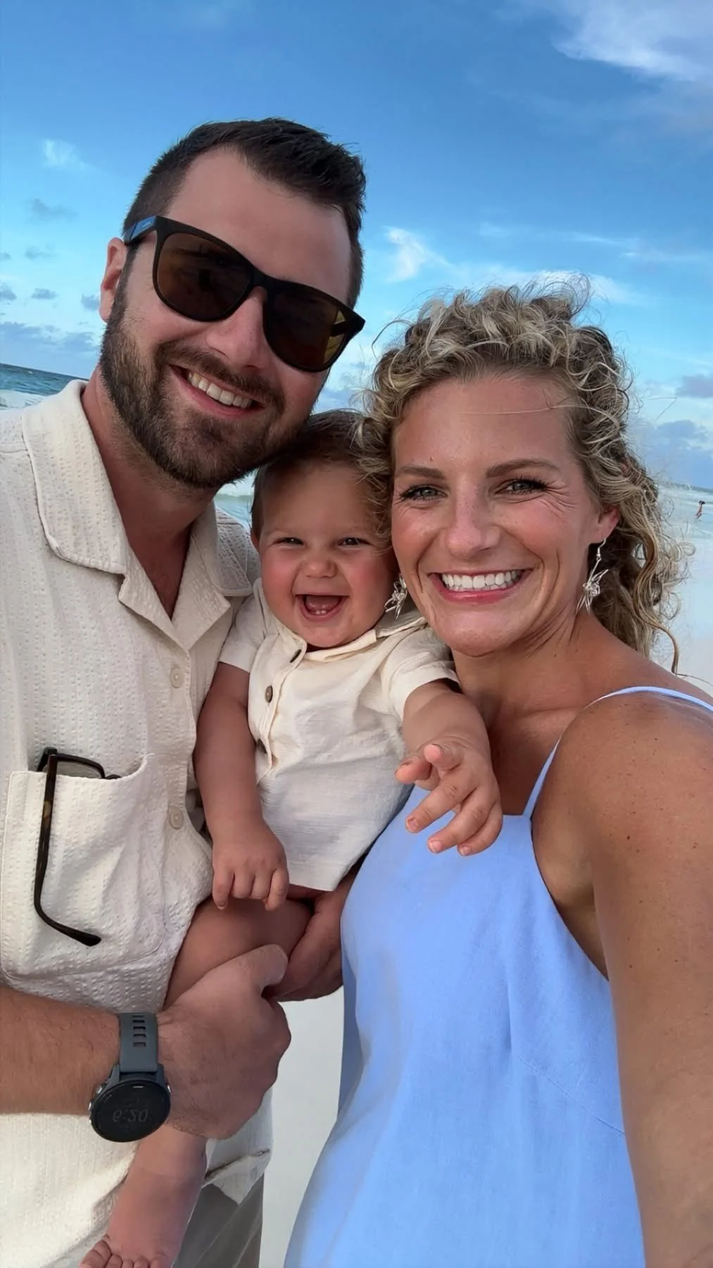 A smiling family of three on the beach with the ocean and blue sky in the background. The man is wearing sunglasses and a light-colored shirt, the woman has curly blonde hair and is wearing a blue dress, and the child is smiling and being held by the man.
