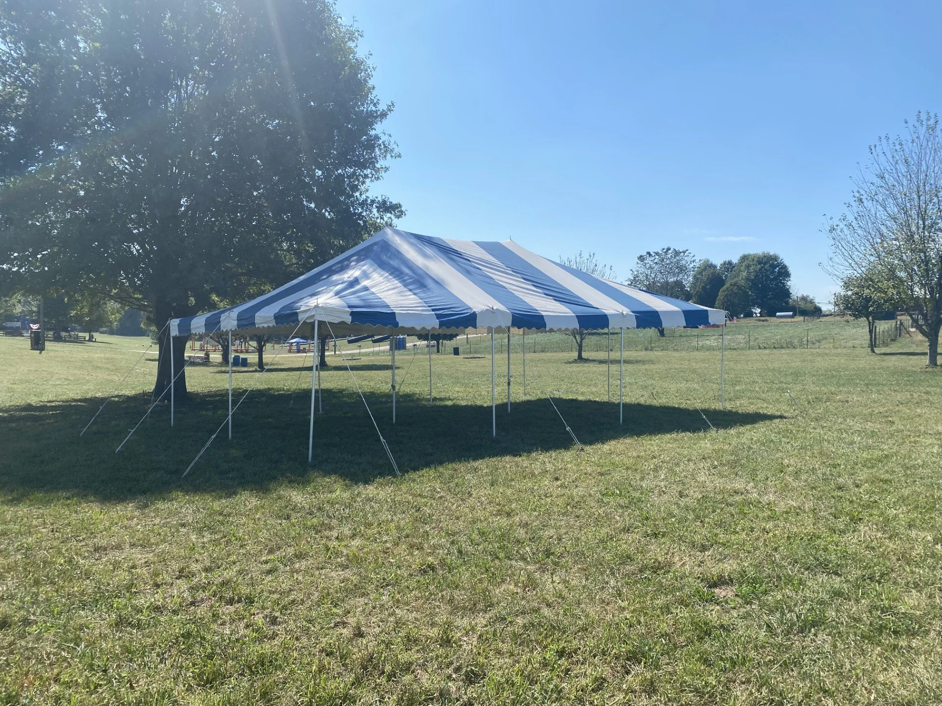 A white and blue striped event tent set up on a grassy field with trees and blue sky in the background.