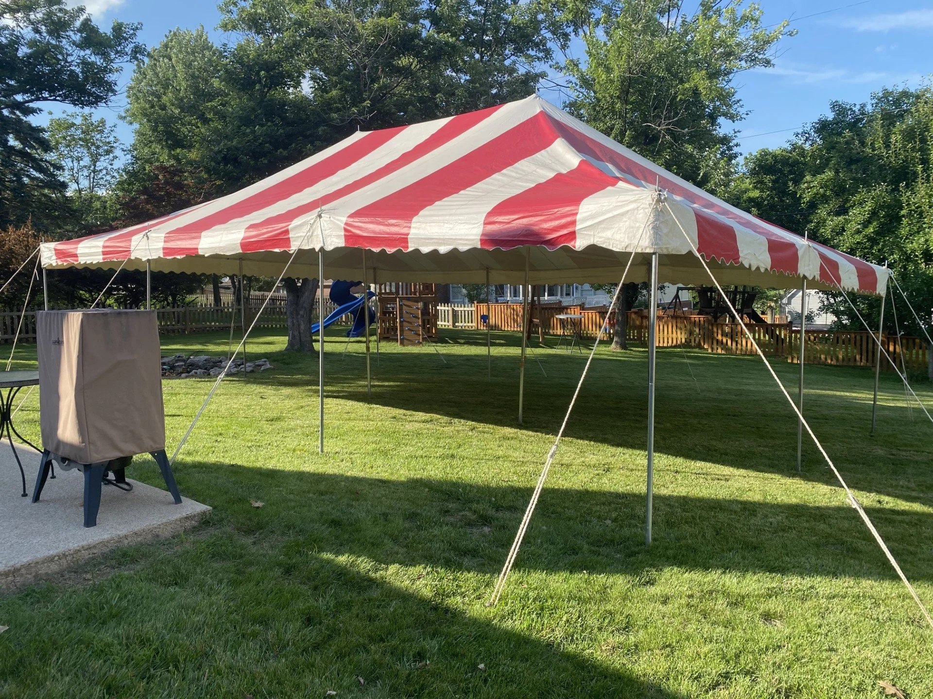 Red and white striped canopy tent set up in a backyard with lush green grass, trees, and a wooden fence, under a partly cloudy sky.