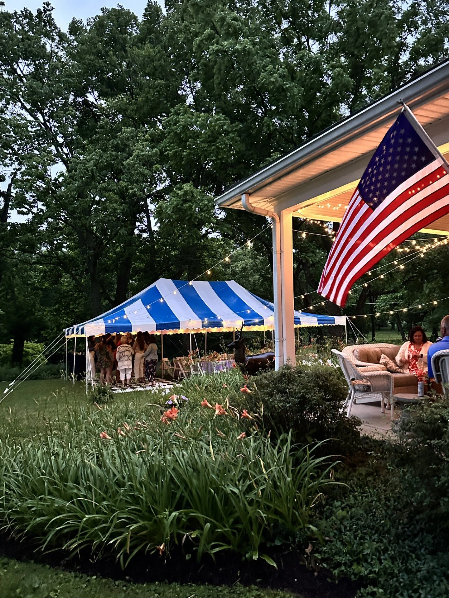 An outdoor gathering with a group of people under a large blue and white striped tent, trees in the background, and an American flag hanging on the porch of a house decorated with string lights, during evening.