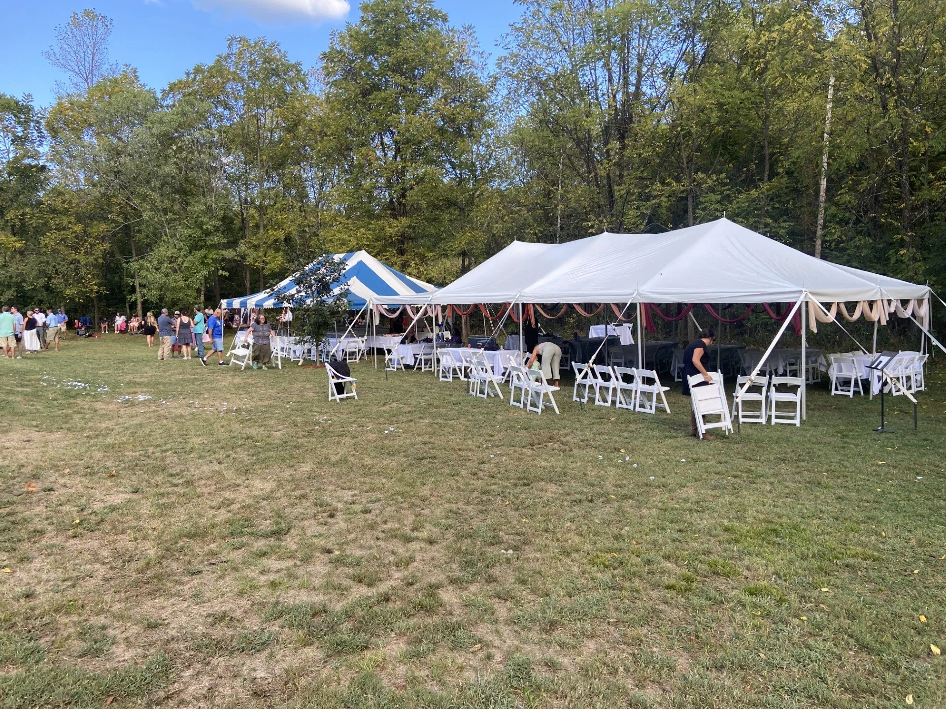 A large outdoor event setup with multiple white tents and chairs, surrounded by a grassy field and dense trees, under a partly cloudy sky.