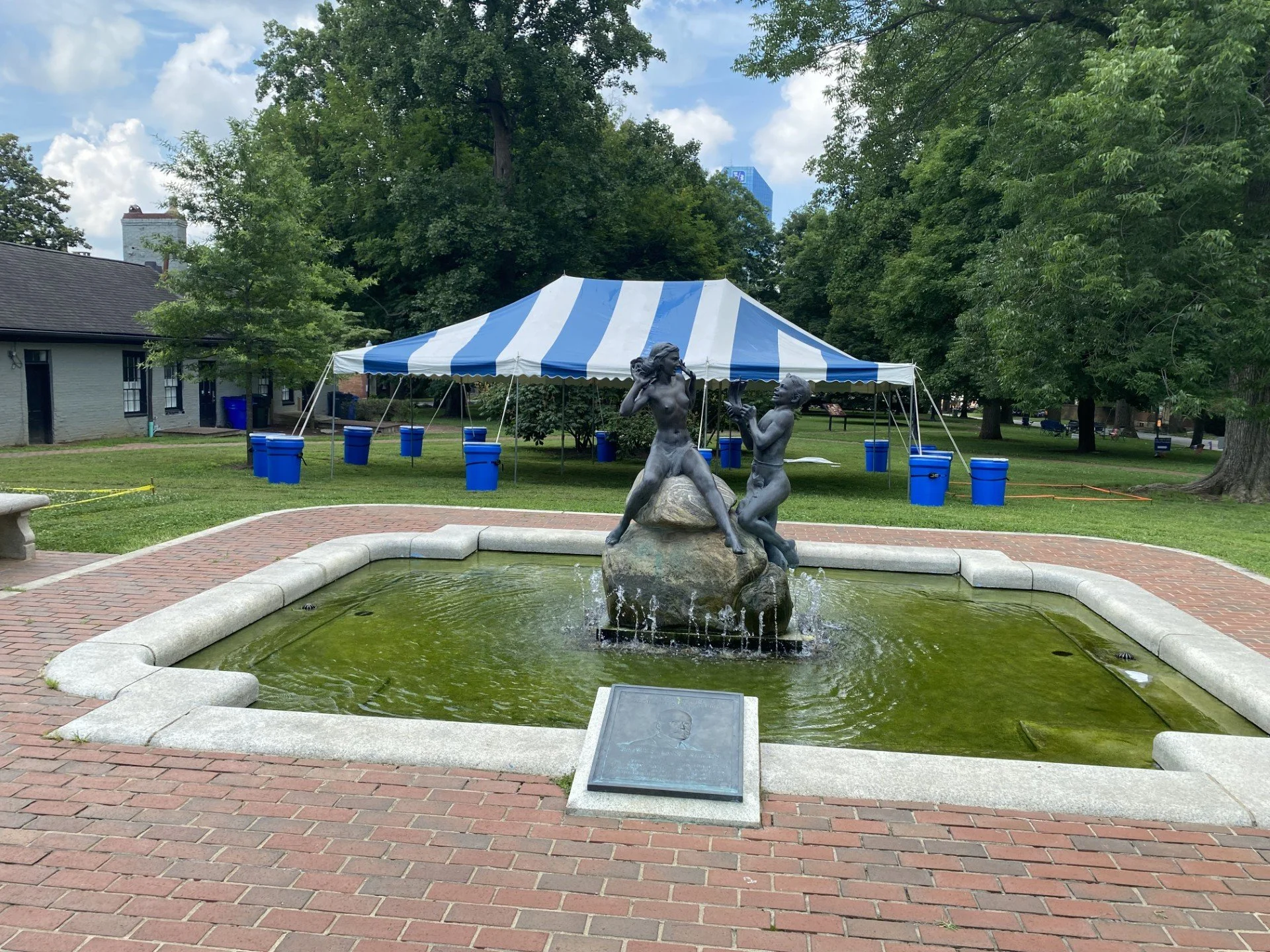 A fountain with a bronze sculpture of two children, one girl and one boy, playing and splashing water in a park. In the background, a blue and white striped tent and green trees are visible.