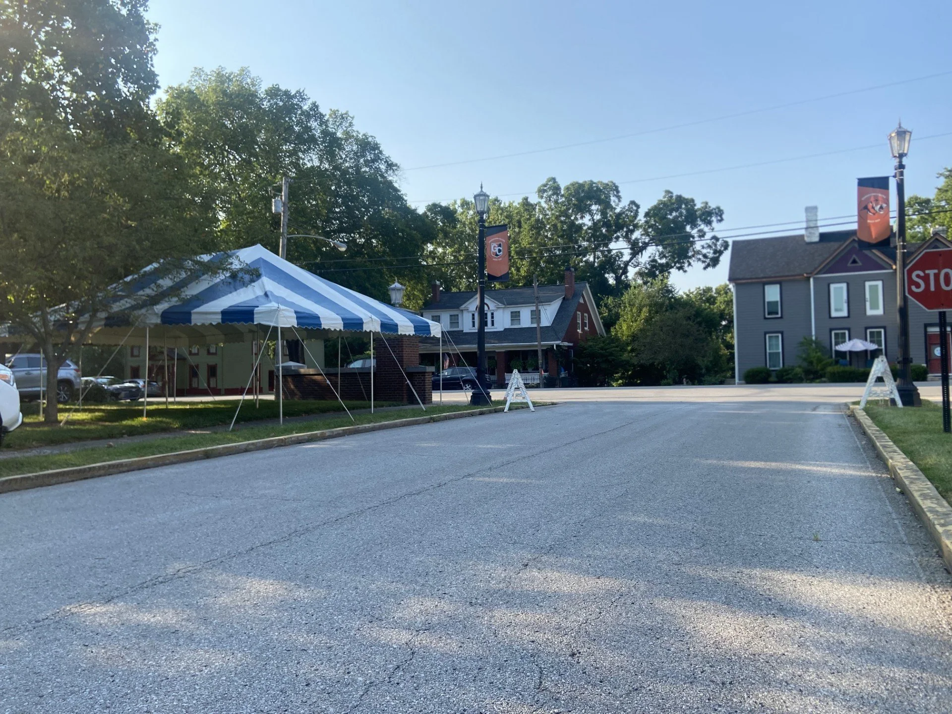 An empty parking lot with a white and blue striped canopy tent on the left. There are houses, trees, and lampposts in the background, with a clear blue sky overhead.