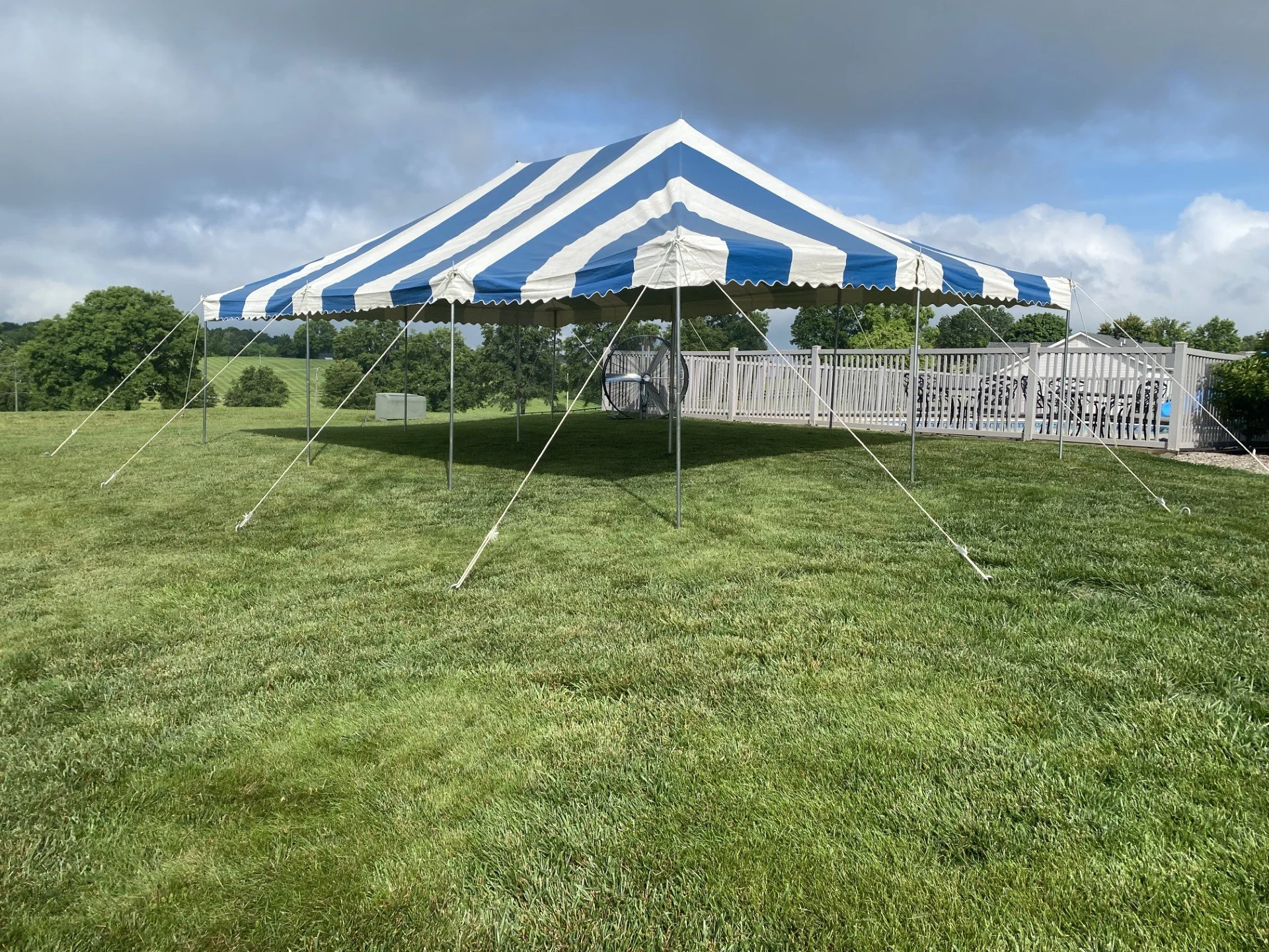 Large outdoor canopy tent with blue and white stripes set up on a grassy area under cloudy sky.