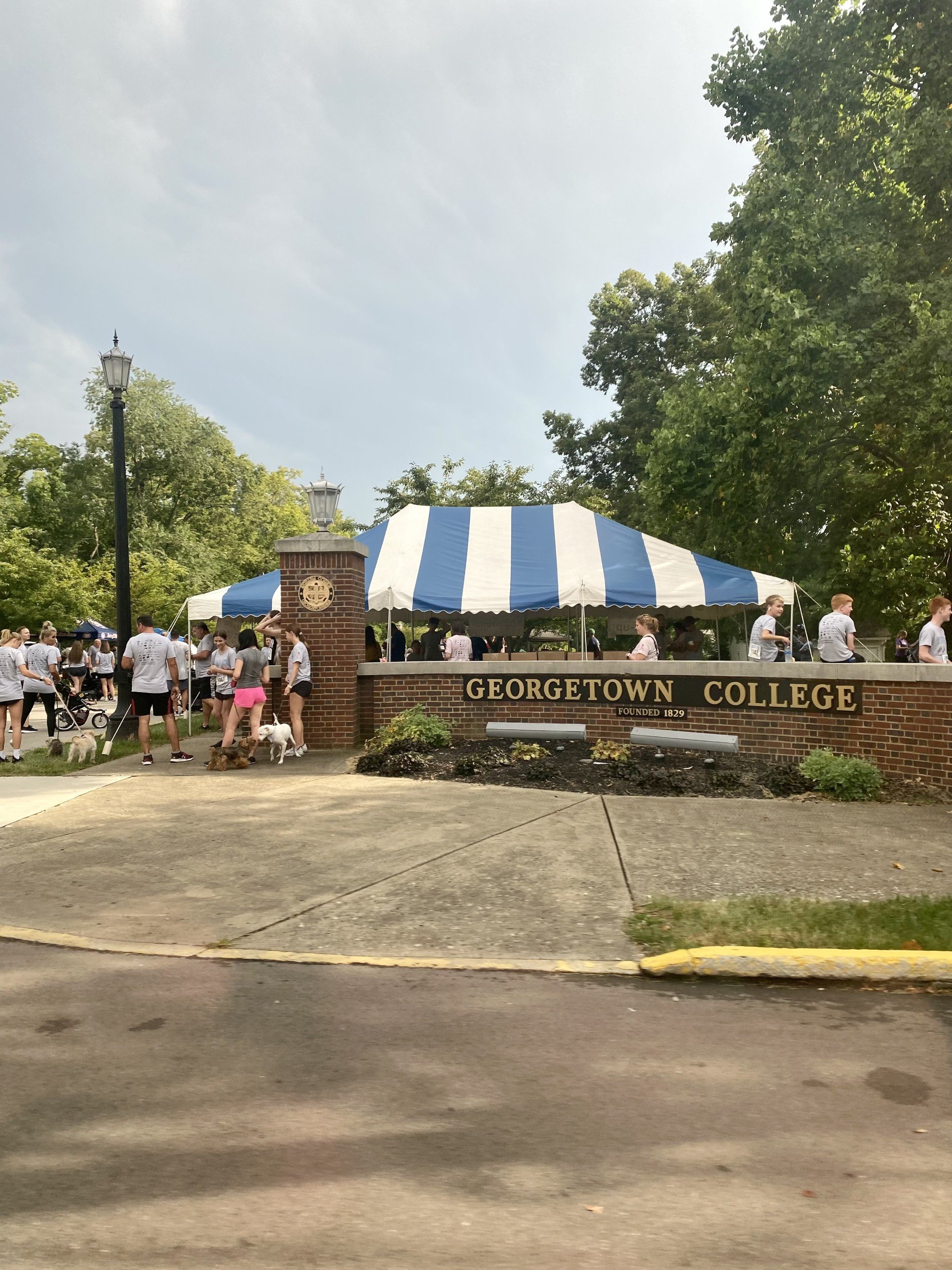 Group of people, some with dogs, gathered near a brick gate with a Georgetown College sign, under a tent with blue and white stripes, in a park-like setting with trees and lamp posts.
