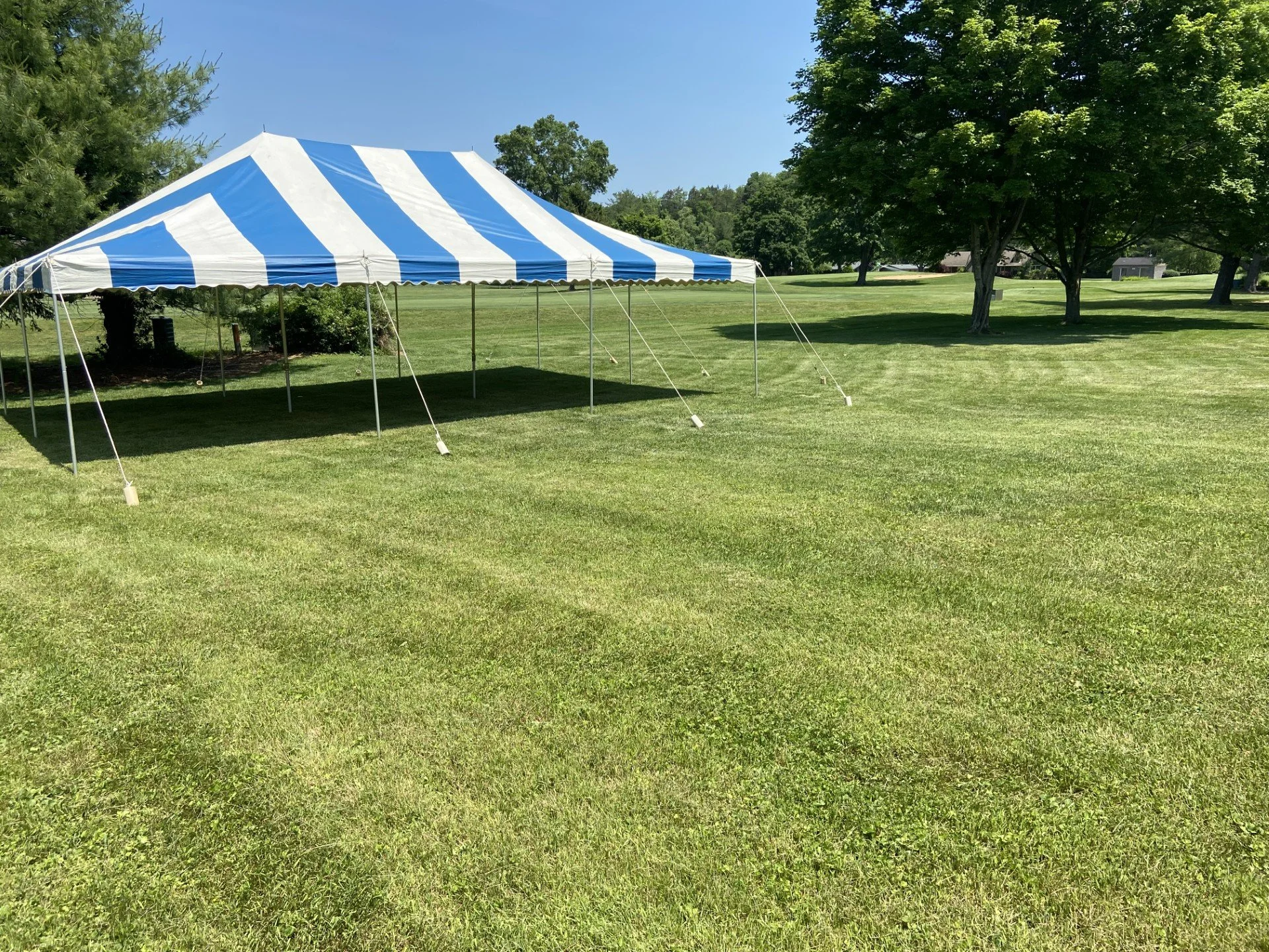 A blue and white striped event tent set up on a grassy lawn, surrounded by trees under a clear blue sky.