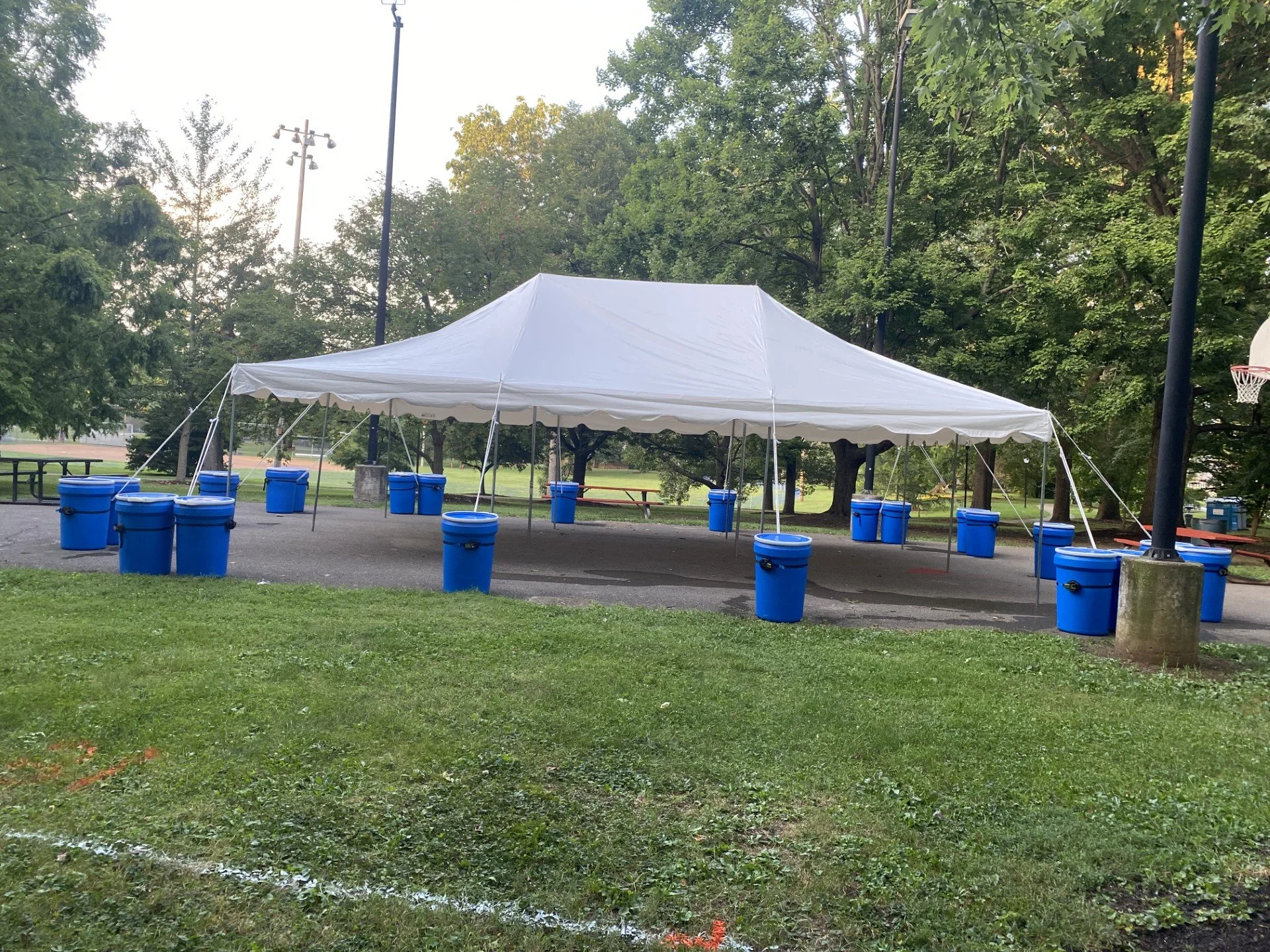 A large white outdoor tent set up in a park with multiple blue water coolers underneath it. The park has green grass and trees, with a basketball hoop on the right and power poles in the background.