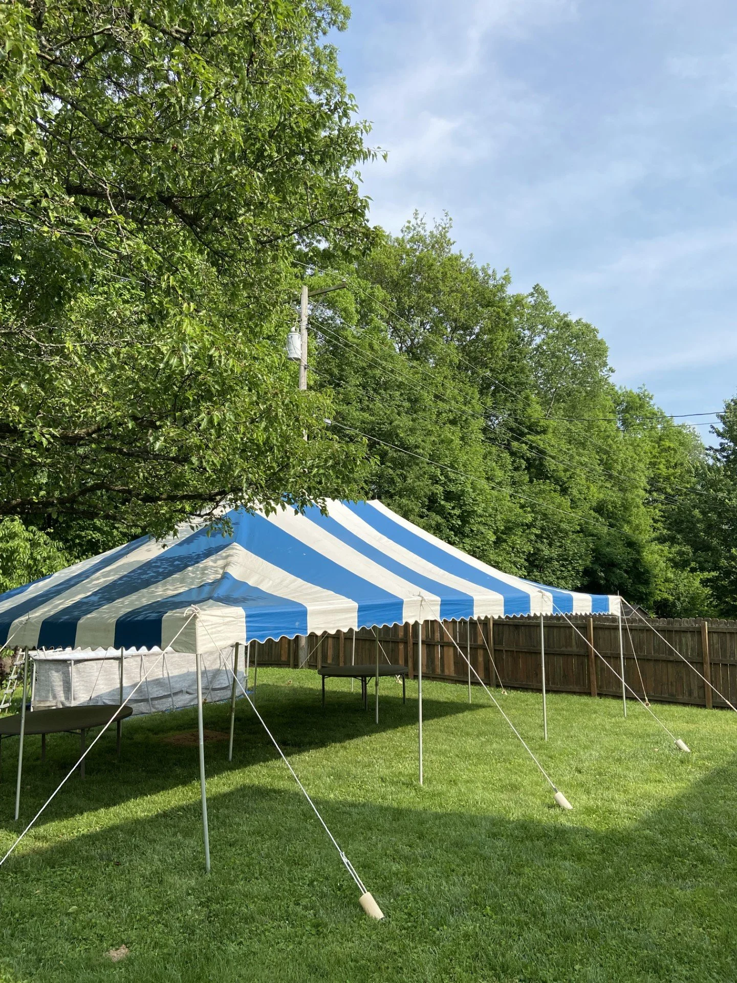 A blue and white striped canopy tent set up on a grassy backyard, with a wooden fence and trees in the background, under a partly cloudy sky.