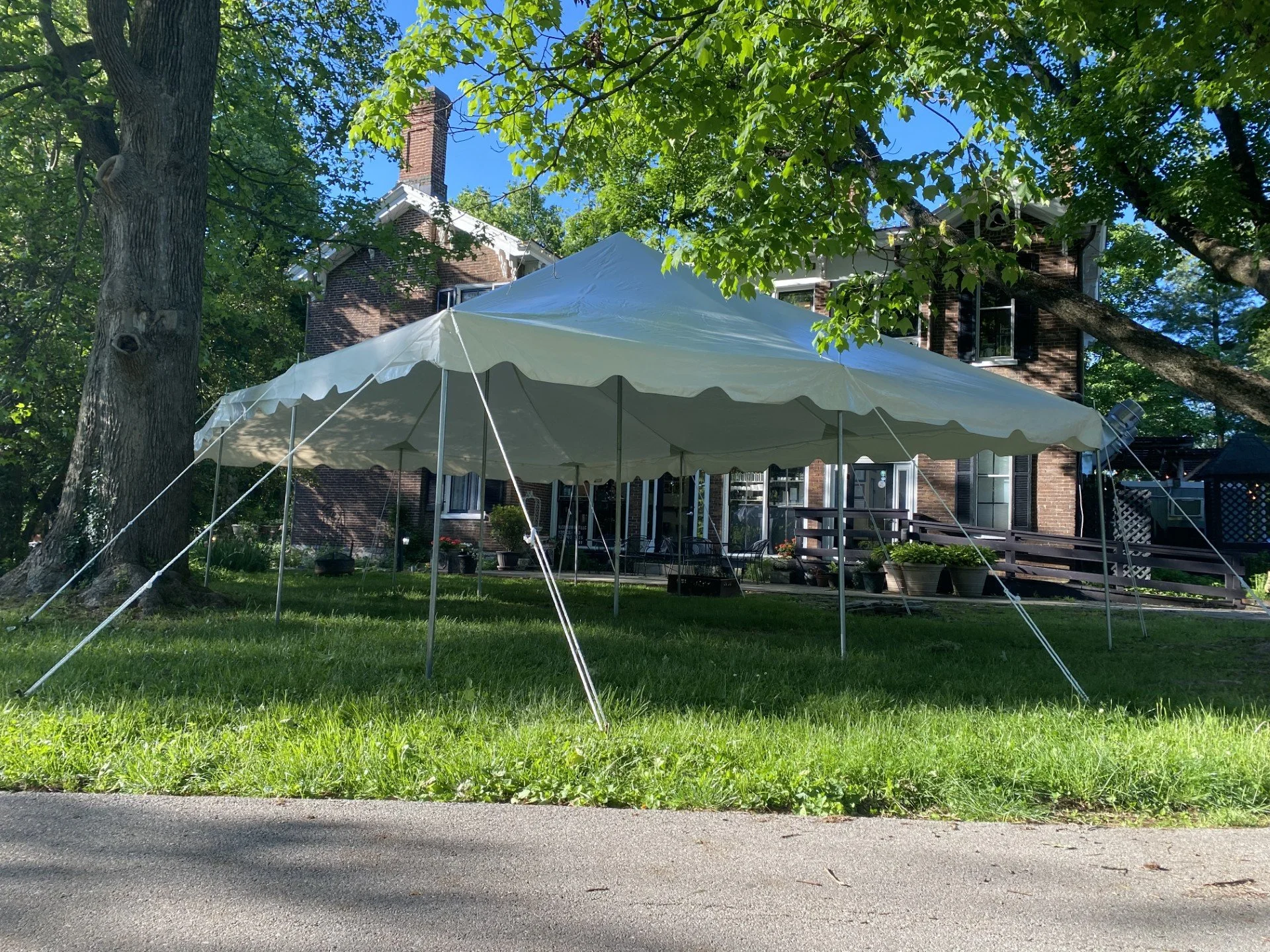 A large white canopy tent set up on a lush green lawn in front of a brick house with open windows. Tall trees with green leaves surround the area, providing shade and a bright blue sky is visible overhead.