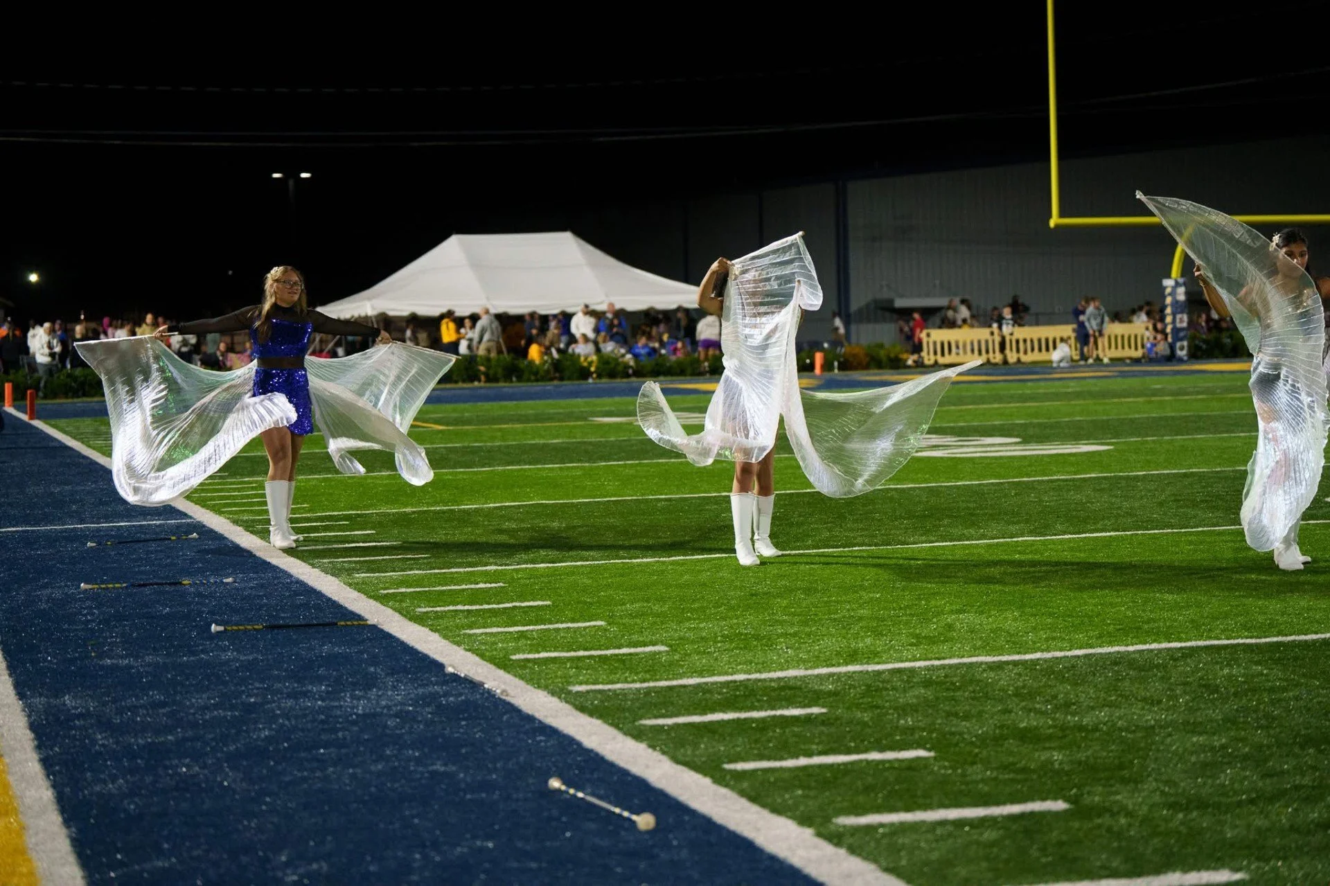 Three performers in white costumes with transparent fabric dancing on a football field at night, with a crowd in the background and a large white tent.