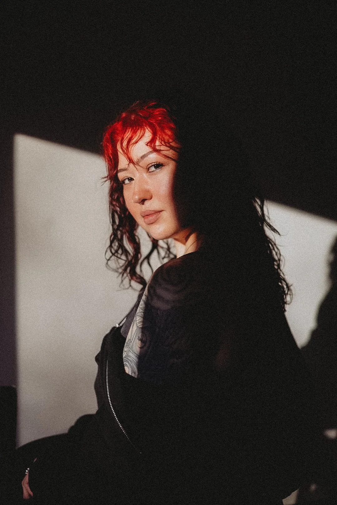 Portrait of a woman with red and black curly hair, wearing a black jacket, looking at the camera with a subtle smile.