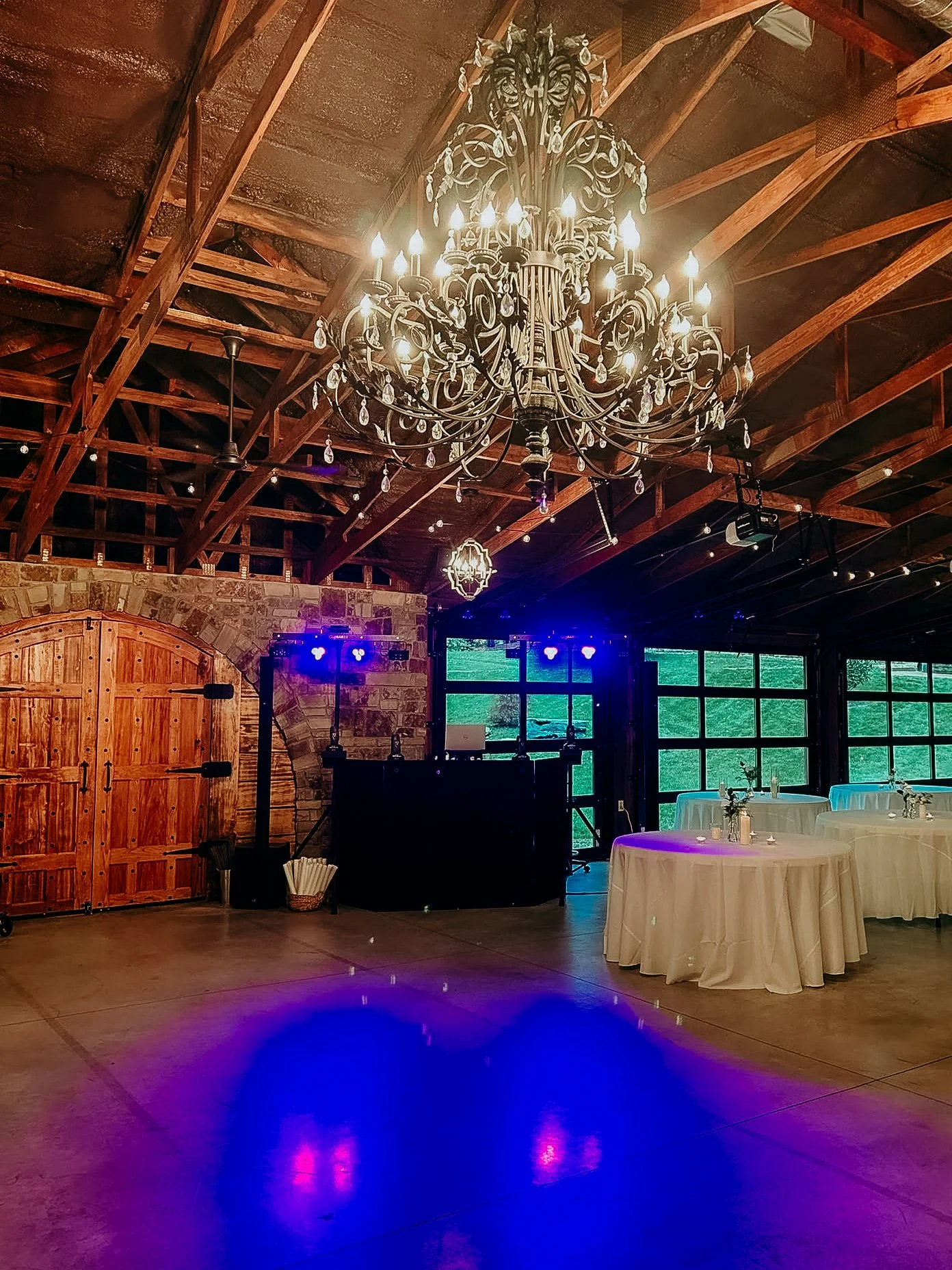 Elegant event space with a large chandelier, round tables with white tablecloths and small floral centerpieces, stone and wood wall, and large windows showing greenery outside, prepared for a celebration or reception.