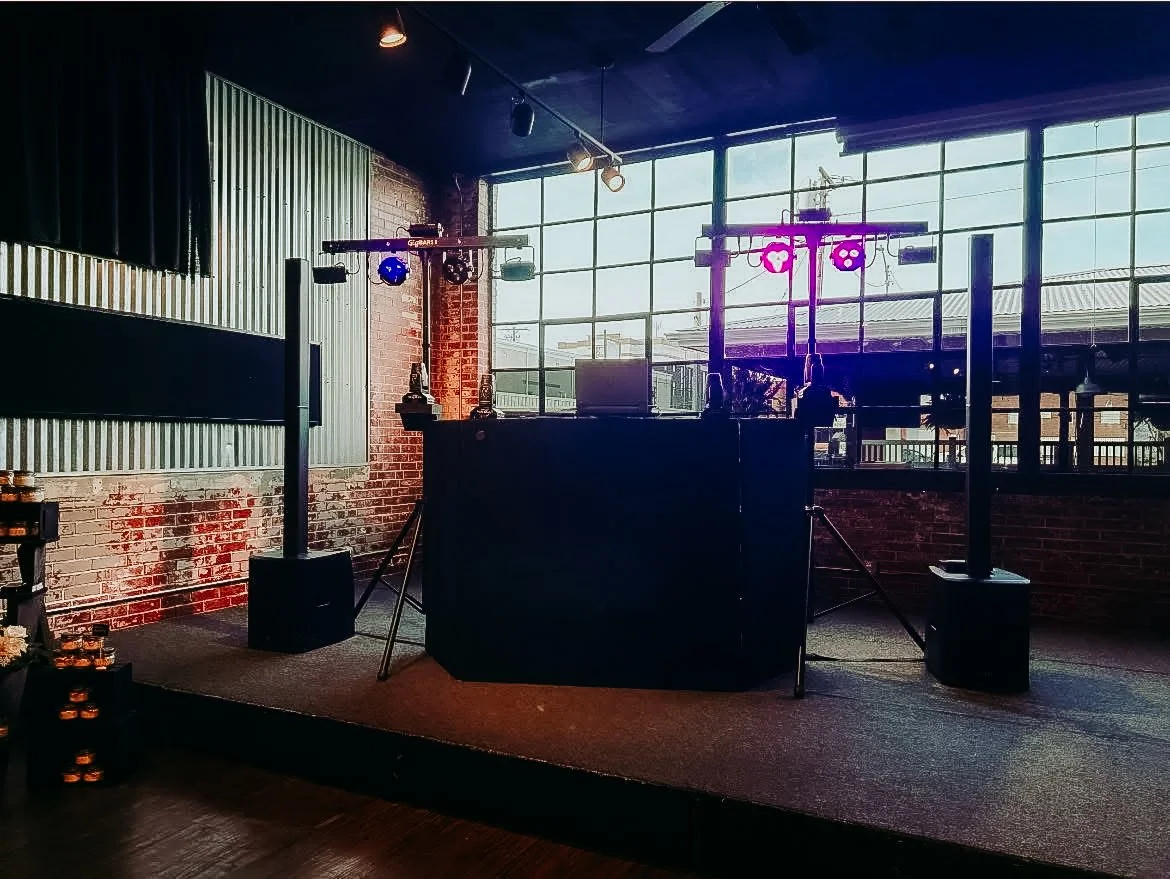 Indoor DJ booth setup with colorful lighting and turntables, brick and metal wall, large window showing city view.