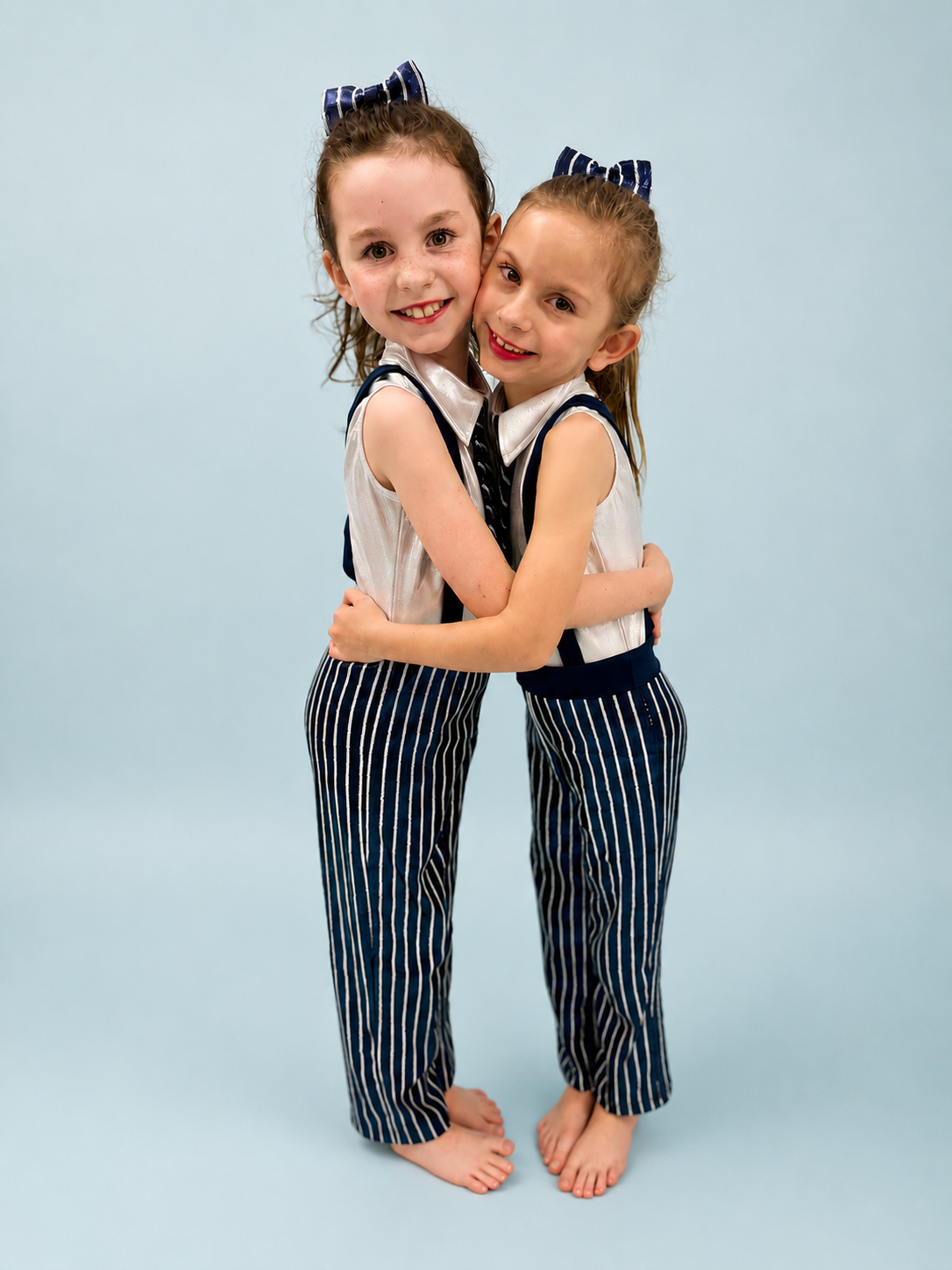 Two young girls in matching striped outfits hugging and smiling at the camera, standing barefoot against a plain light blue background.