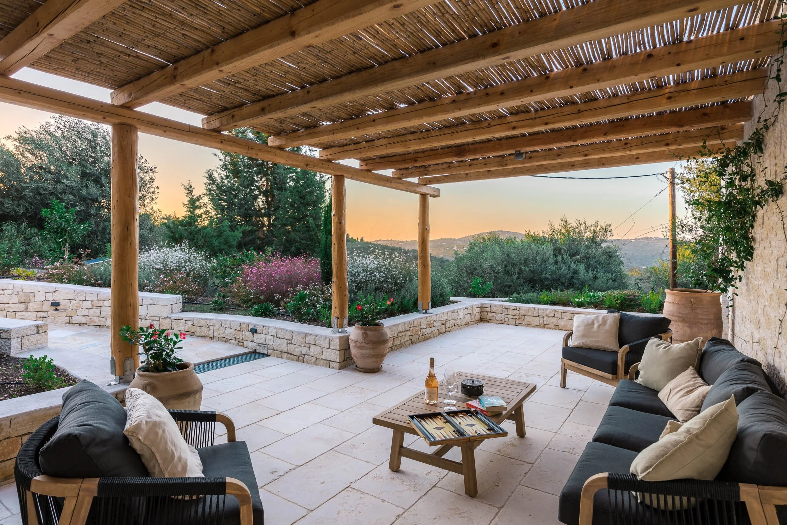 Outdoor patio with stone flooring and wooden roof, surrounded by garden with flowering plants, stone wall, and scenic hills in the background. Contains black cushioned seating with beige pillows, and a small wooden coffee table with a bottle of wine,