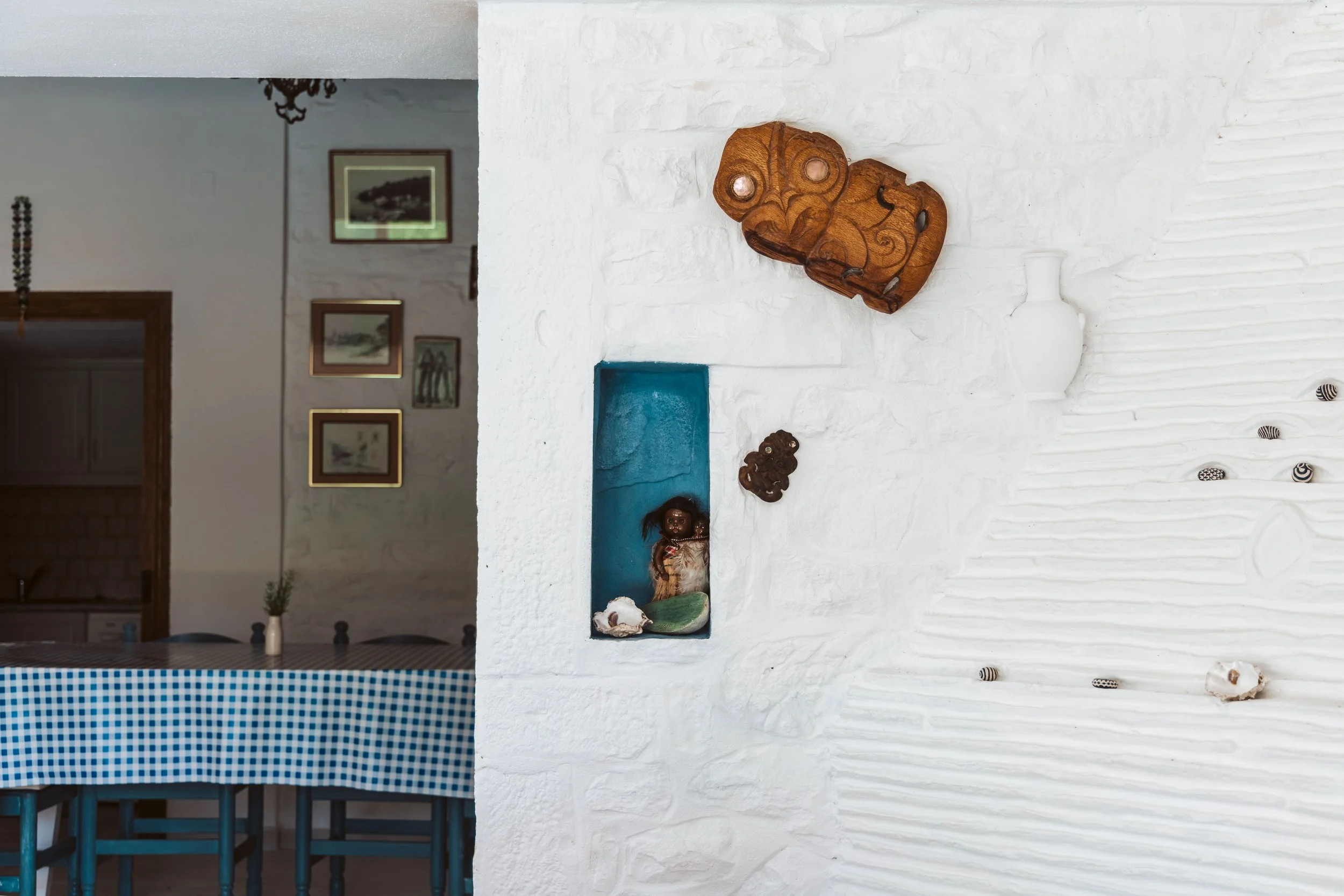 Interior wall with rustic decorations, including a carved wooden mask, a white vase, seashells, and small decorative items, with a gallery of framed pictures visible in the adjacent room.