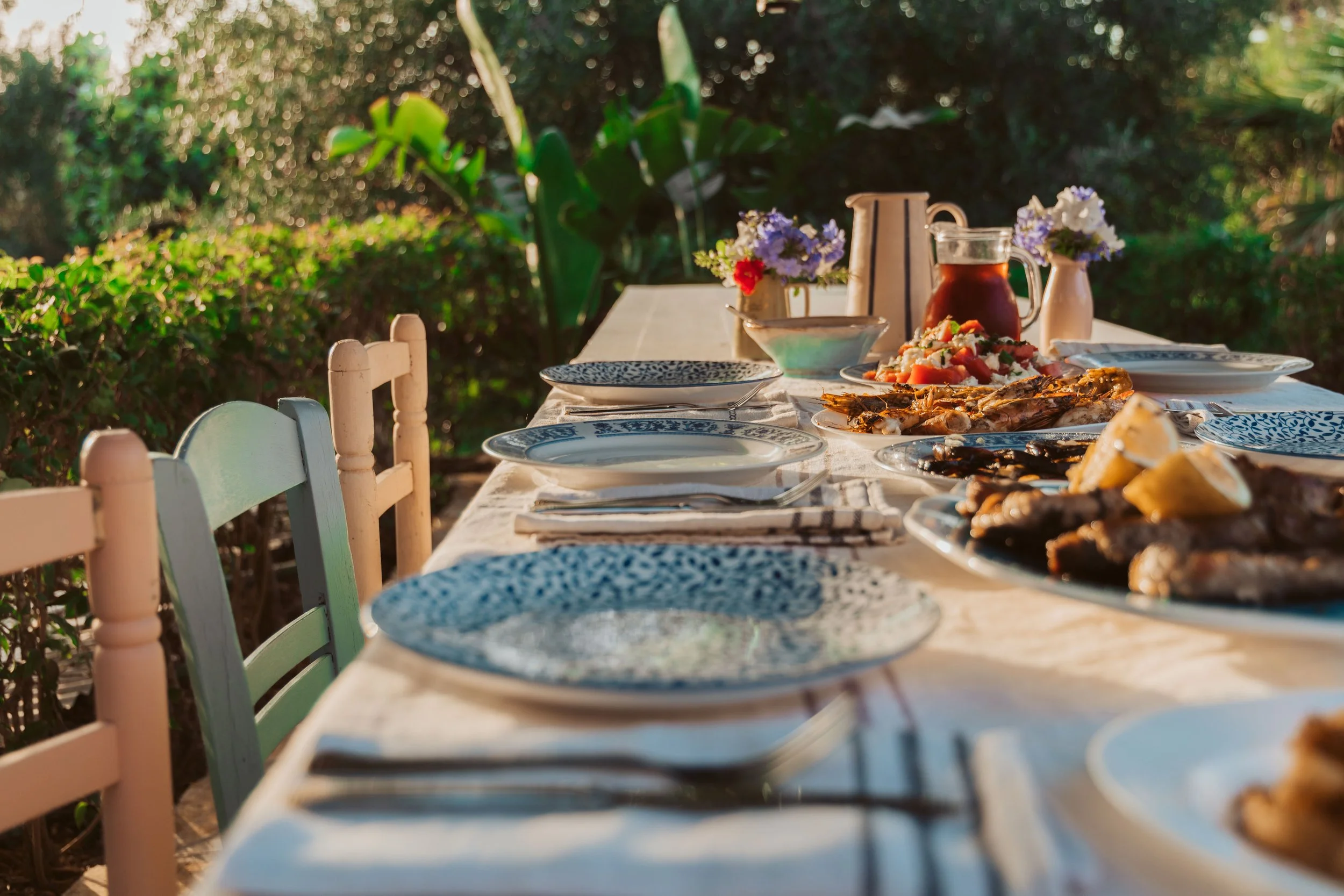 A table set for outdoor meal with plates of grilled meats, vegetables, sauces, and a pitcher of iced tea, surrounded by pastel chairs and decorated with small flower arrangements.