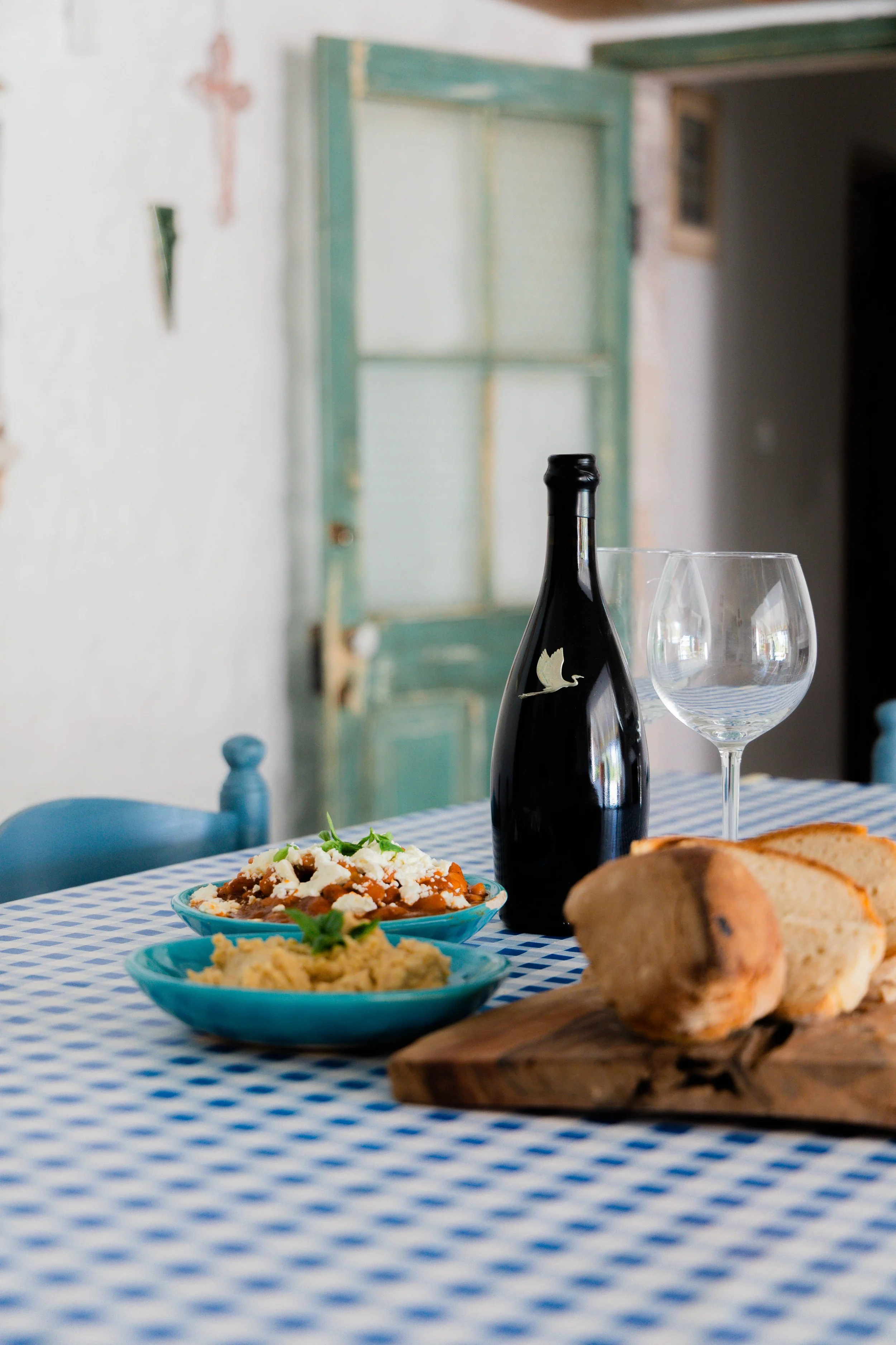 A dining table with a blue and white checkered tablecloth set with two blue bowls of food, a bottle of wine with a bird silhouette on it, two empty wine glasses, and slices of bread on a wooden cutting board, in a rustic room with a distressed turquo