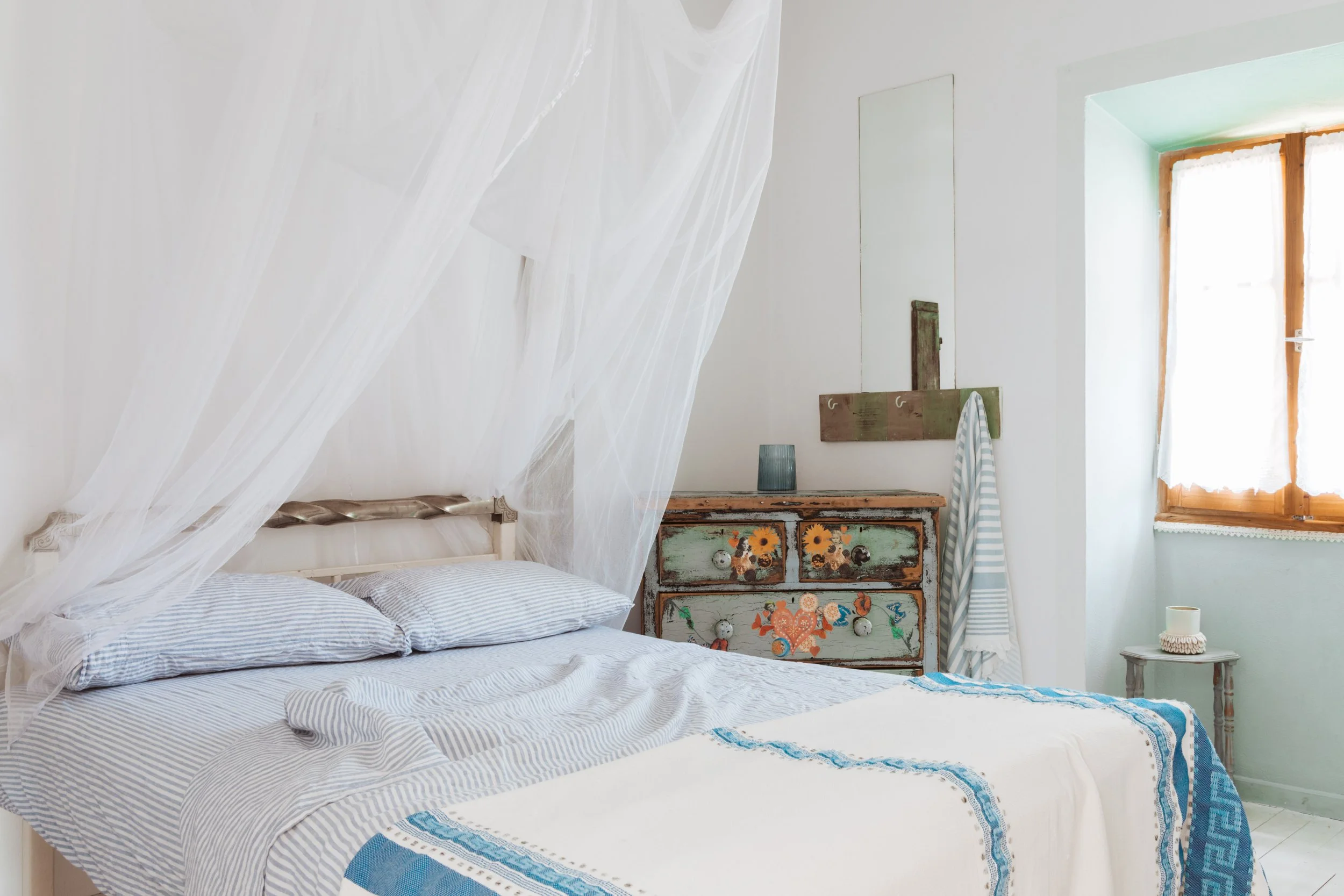 A cozy bedroom with a white bed featuring striped pillows and a white and blue blanket. A sheer canopy drapes over the bed. A vintage painted wooden dresser with floral artwork stands against the wall, with a small blue lamp on top. An ironing board 