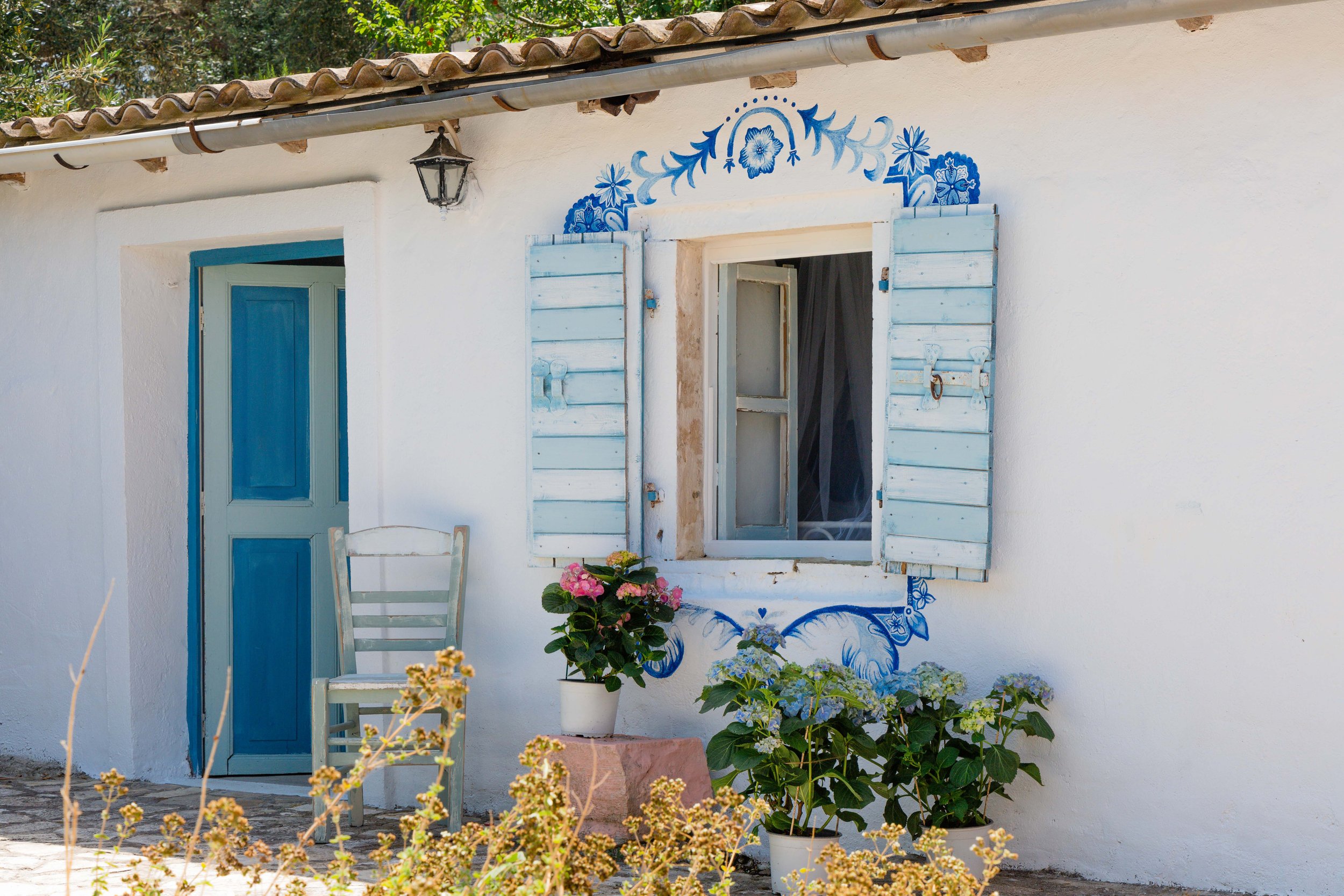 A white house with blue accents, including shutters and door, with decorative blue floral artwork above the window. There is a small chair and potted plants outside, and the house has a tiled roof and an outdoor lantern.