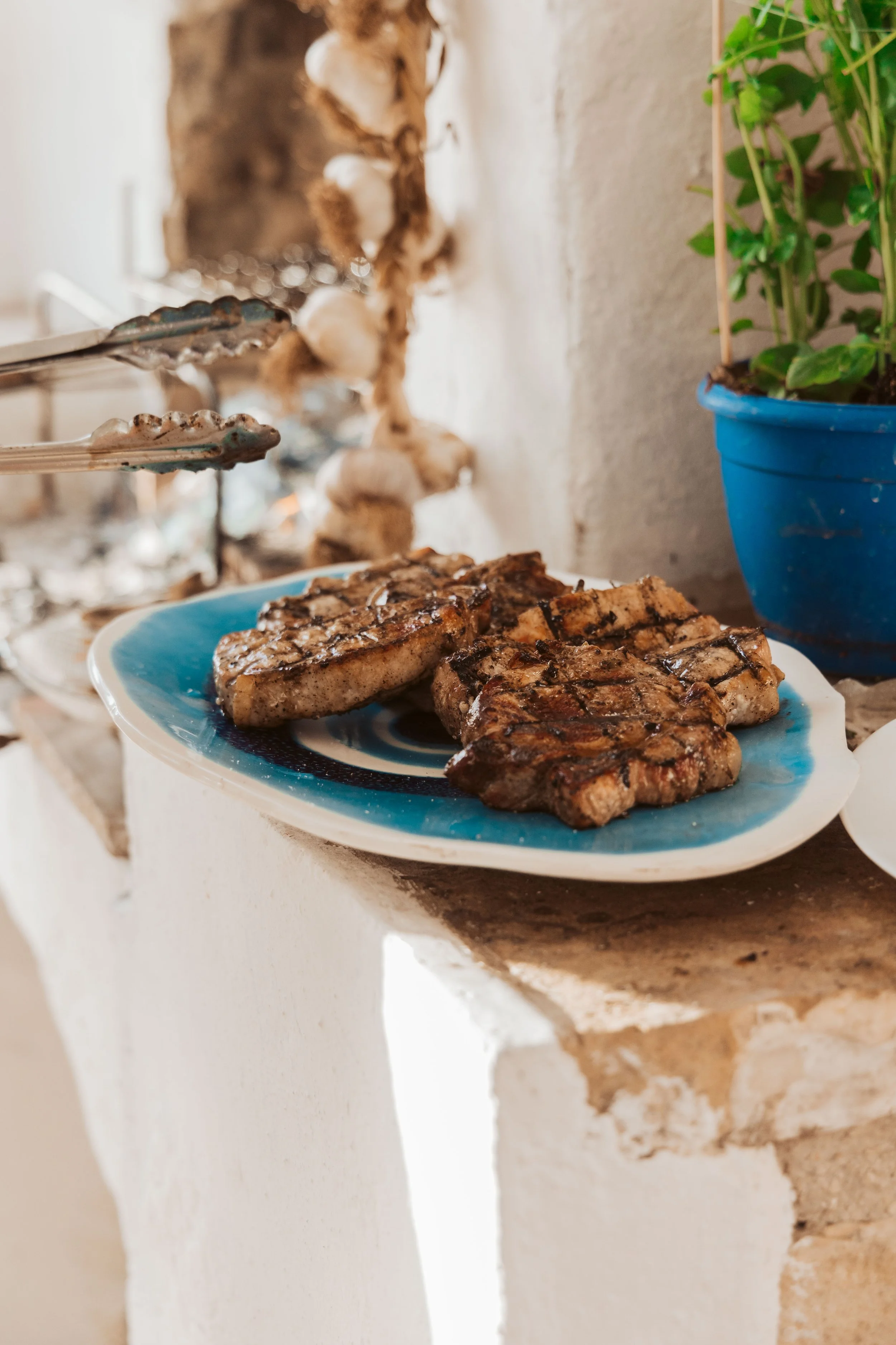 Grilled meat chorizo on a blue and white ceramic plate on a stone ledge, with tongs nearby and a potted green plant in a blue pot in the background.
