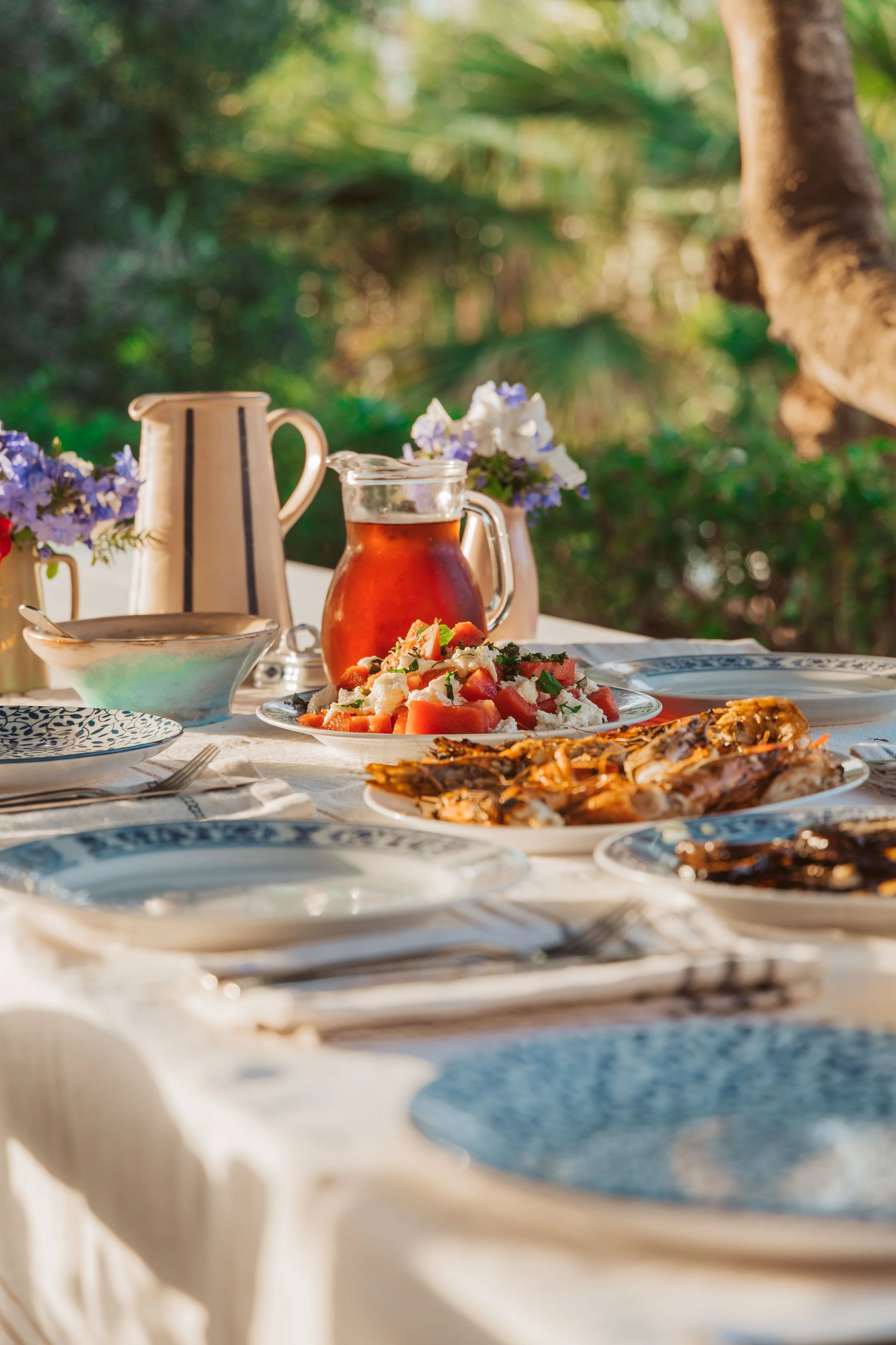 An outdoor table set with plates of food, a pitcher of tea, and vases of flowers, with a green garden in the background.