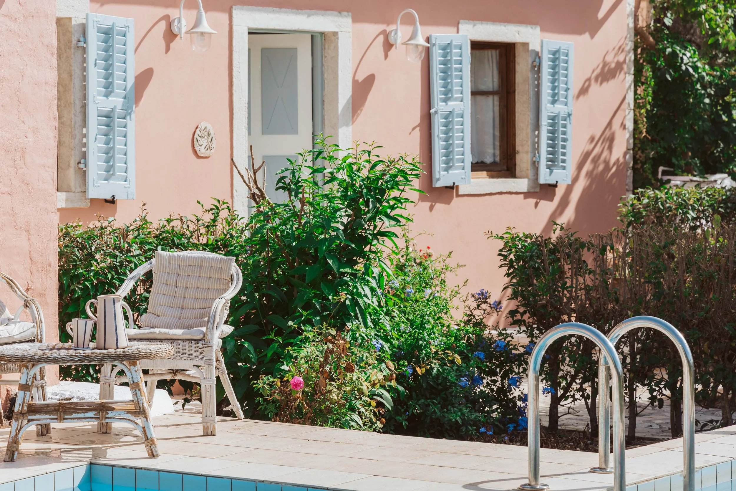 A pink house with white shutters, a small pool with a metal ladder, wicker chairs, a small table with cups, and lush green bushes and flowers.