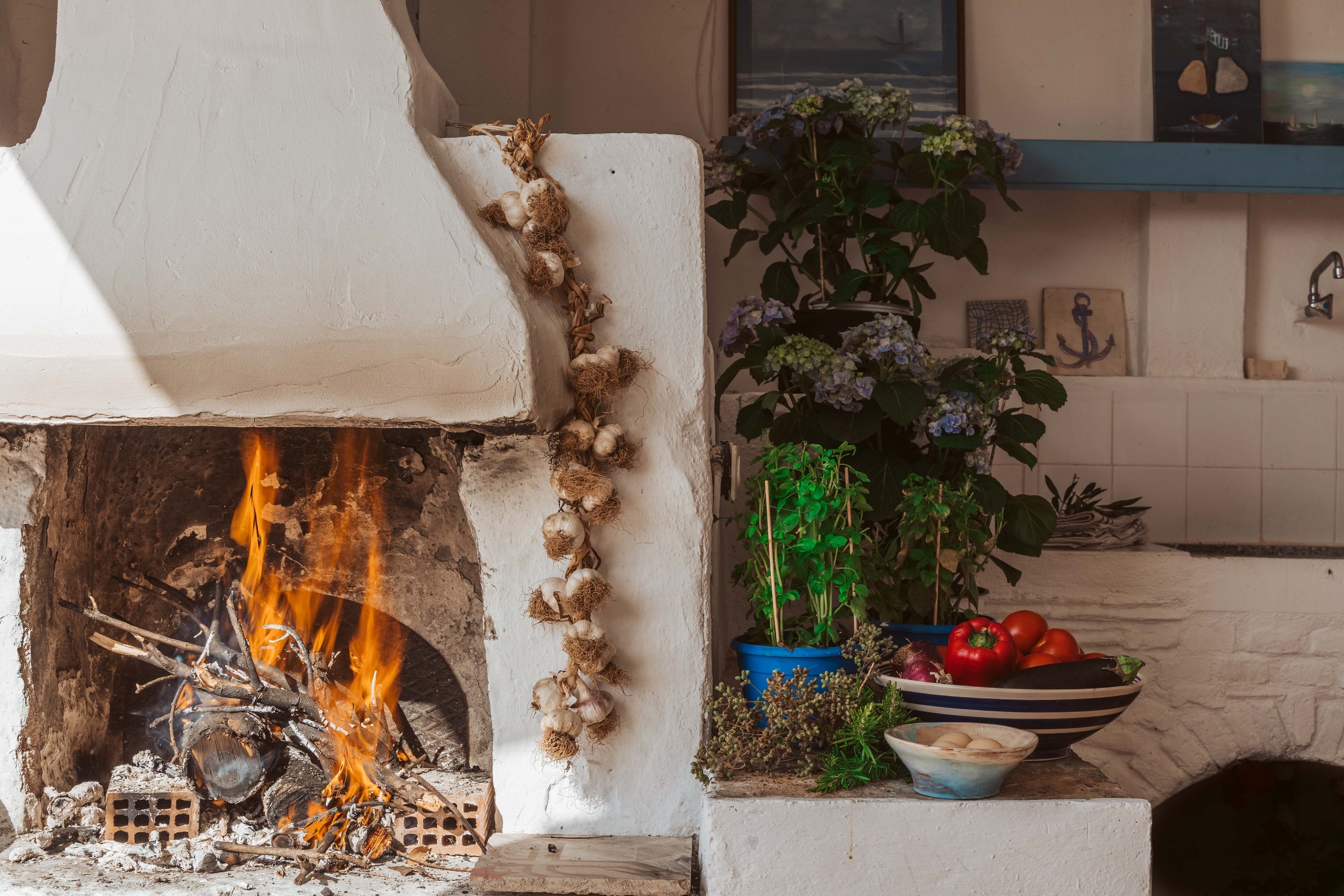 Fire burning in a rustic white fireplace, decorated with a string of garlic and herbs hanging on the side. To the right, there are potted plants, fresh vegetables including tomatoes, bell peppers, and eggplant, as well as a bowl with garlic cloves. T