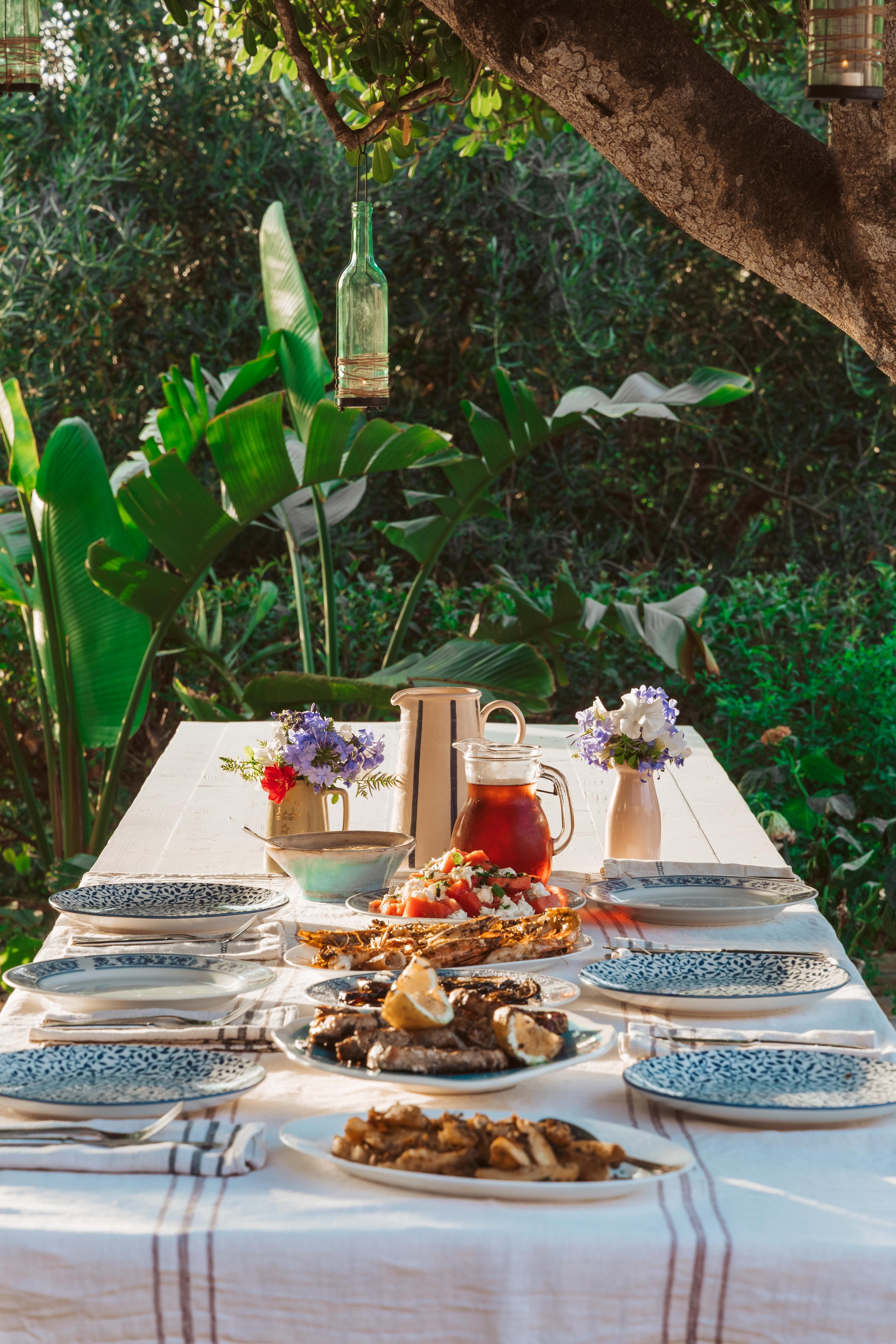 Outdoor dining table set with plates, bowls, dishes of food, two flower vases with purple and white flowers, a pitcher of tea, a teapot, and a bottle, under a tree with green foliage.
