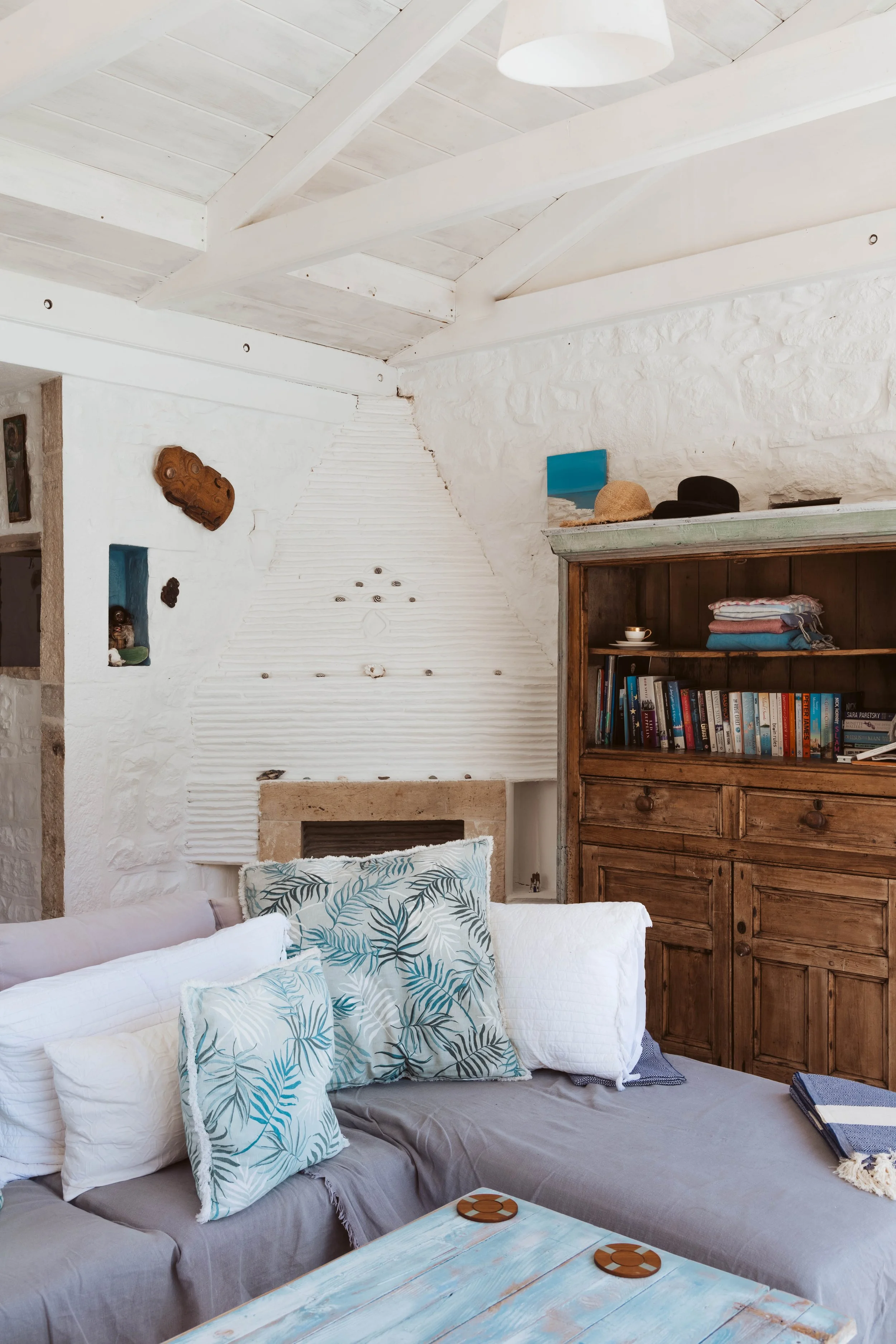 Cozy living room with white walls, wooden ceiling beams, a navy sectional sofa with tropical leaf pillows, a distressed blue coffee table with coasters, and a wooden bookshelf filled with books and folded towels.