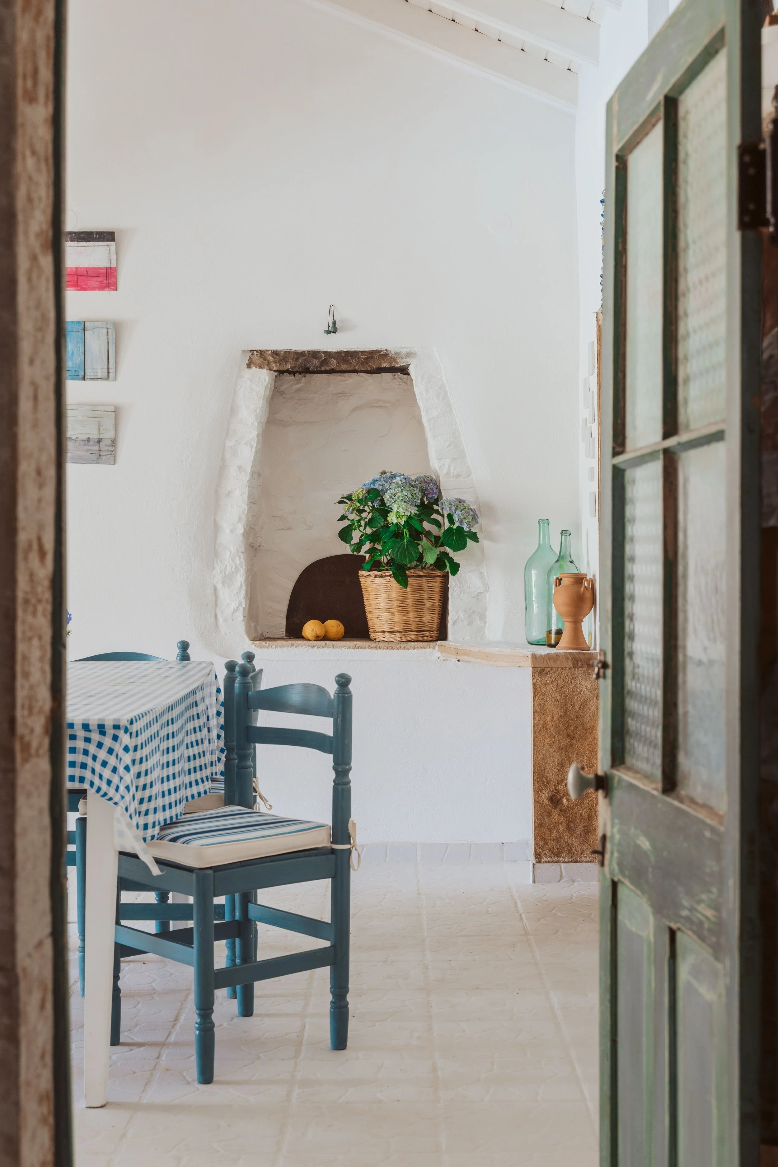 Interior of a cozy room with a white wall, blue checkered tablecloth on a table, blue wooden chair, decorative shelf with paintings, a floral arrangement in a basket, and glass bottles on a ledge. Wooden door partially open.