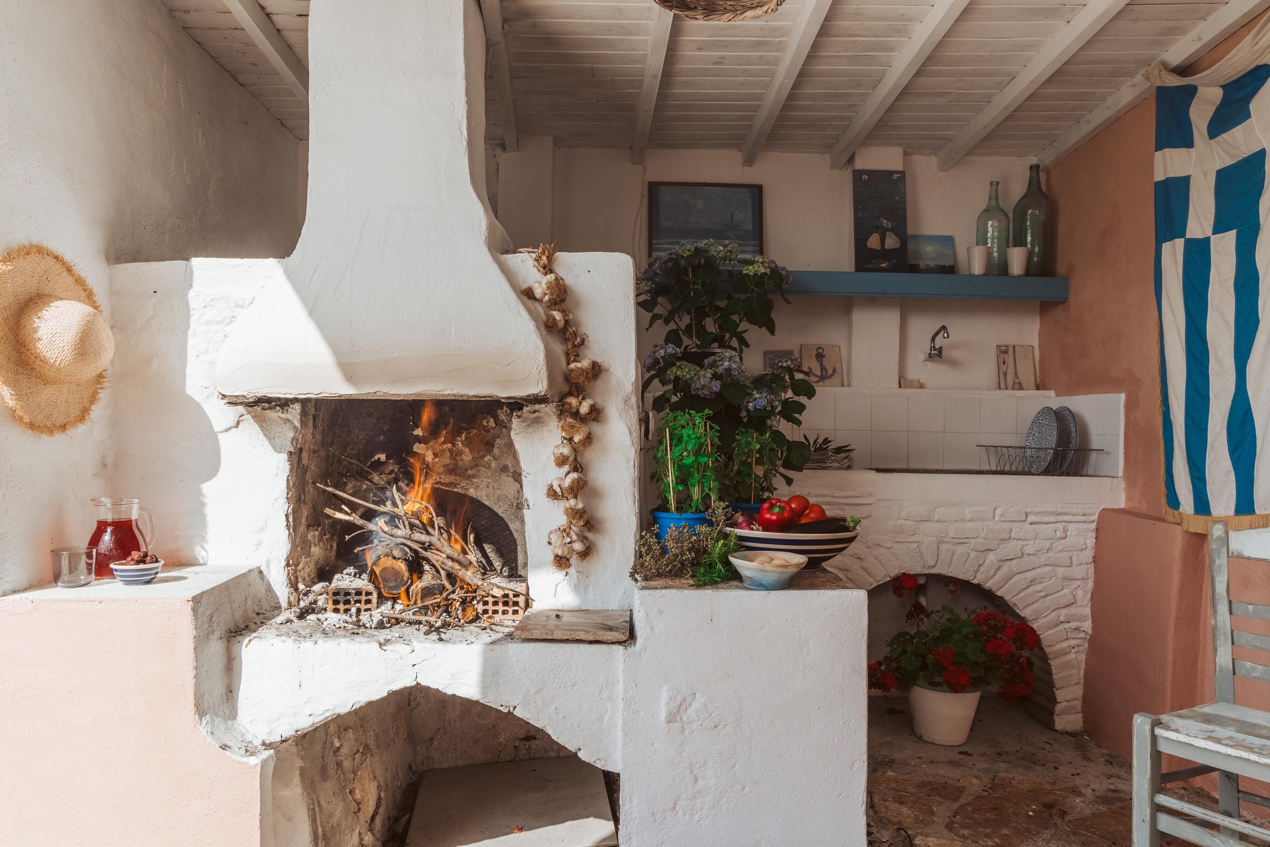 Cozy indoor fireplace with a burning fire, decorated with garlic strings, surrounded by ceramic and glass jars, bowls of fruit, plants, and paintings on the wall in a rustic styled room.