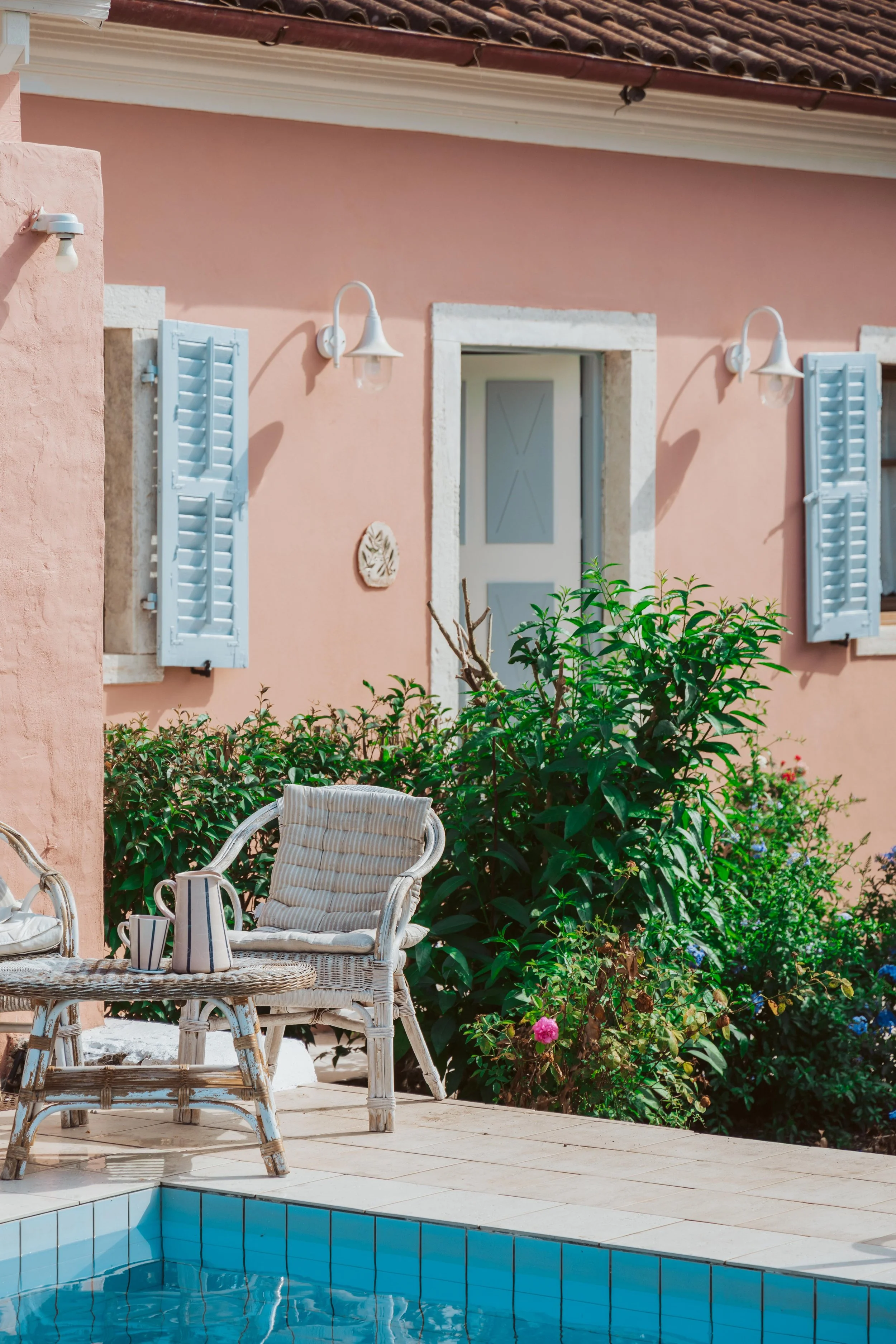 A pink house with white trim and light blue window shutters, outdoor wall lamps, a patio with wicker chairs, a small table with a pitcher and a cup, a pool with blue tiles, and lush green shrubs with flowers.