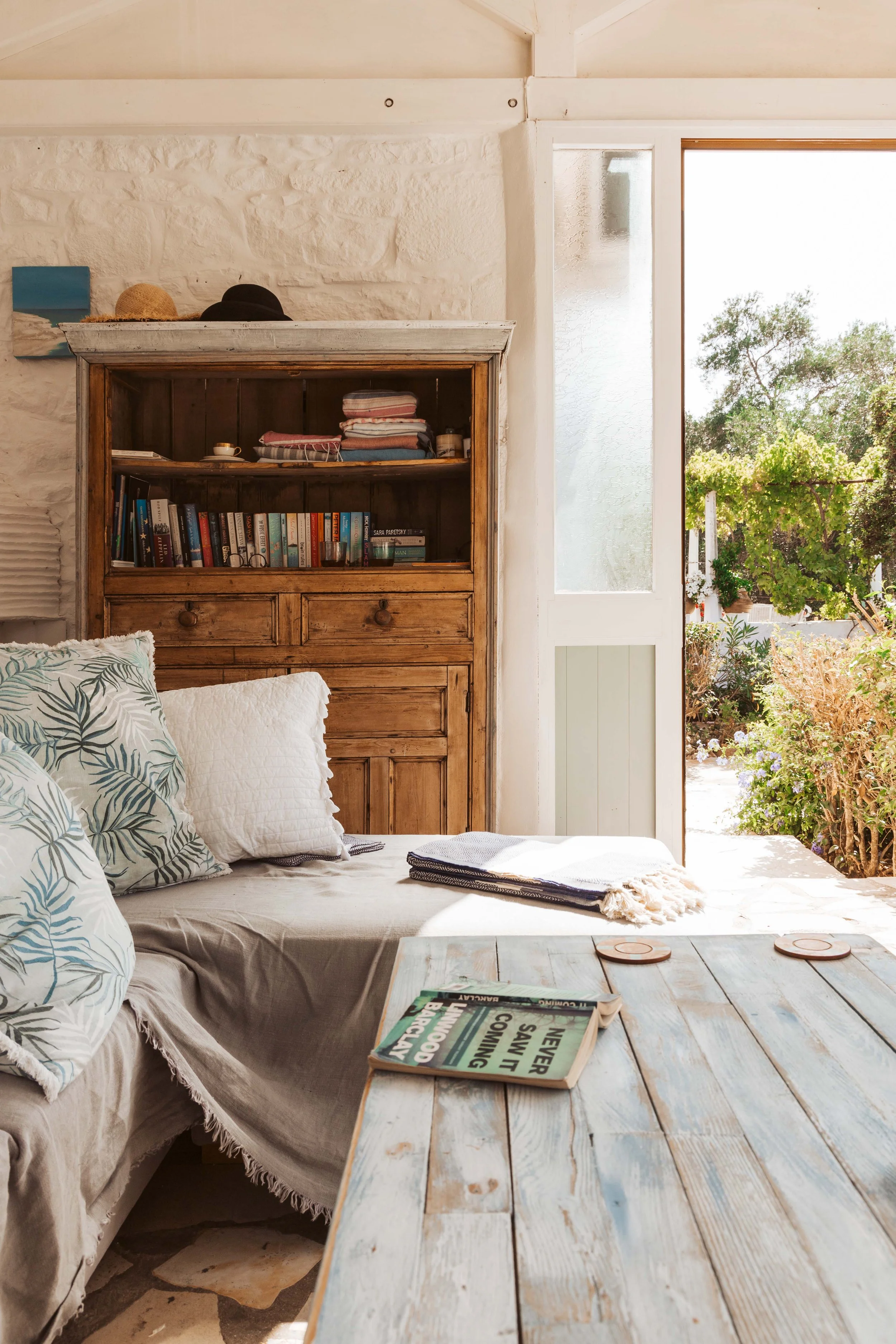 Cozy living room with a couch, decorative pillows, a rustic wooden bookcase filled with books, and an open door leading outside to a garden with greenery and sunlight.