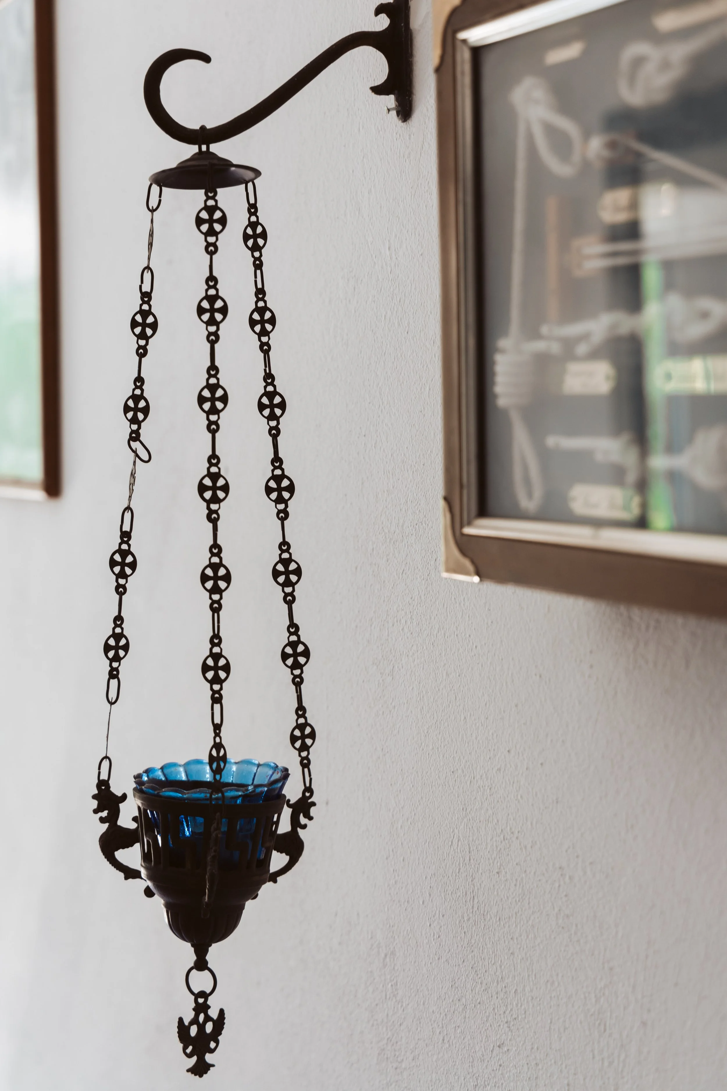 A hanging metal wind chime with chains and a blue glass bowl at its center, mounted on a white wall next to a framed display case.