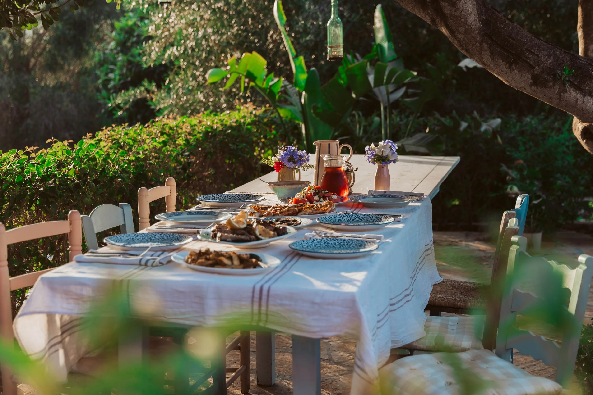 Outdoor dining table set with plates, food, and flowers, surrounded by greenery in sunlight