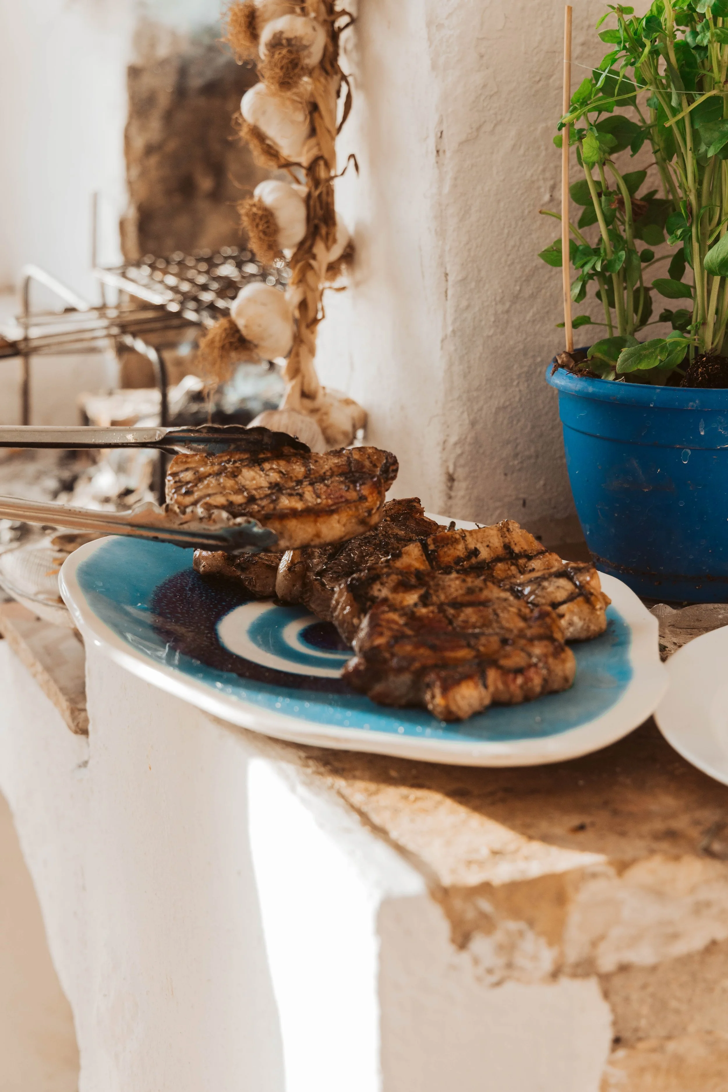 Grilled steaks on a decorative blue and white plate with tongs resting on top, next to a blue pot with a green plant, behind a rustic stone and plaster surface.